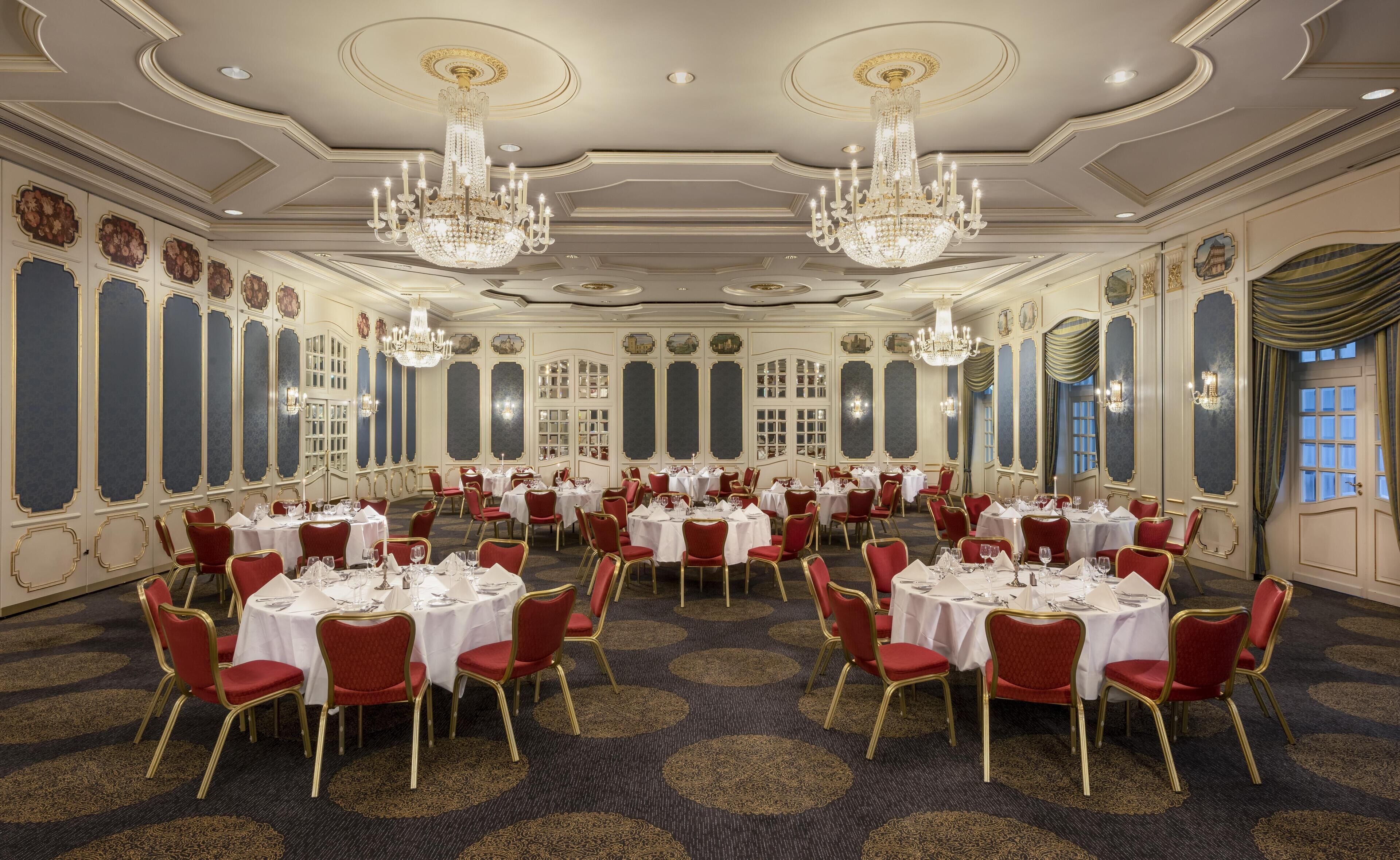 A banquet hall with round tables, red chairs, chandeliers, and ornate wall decorations.