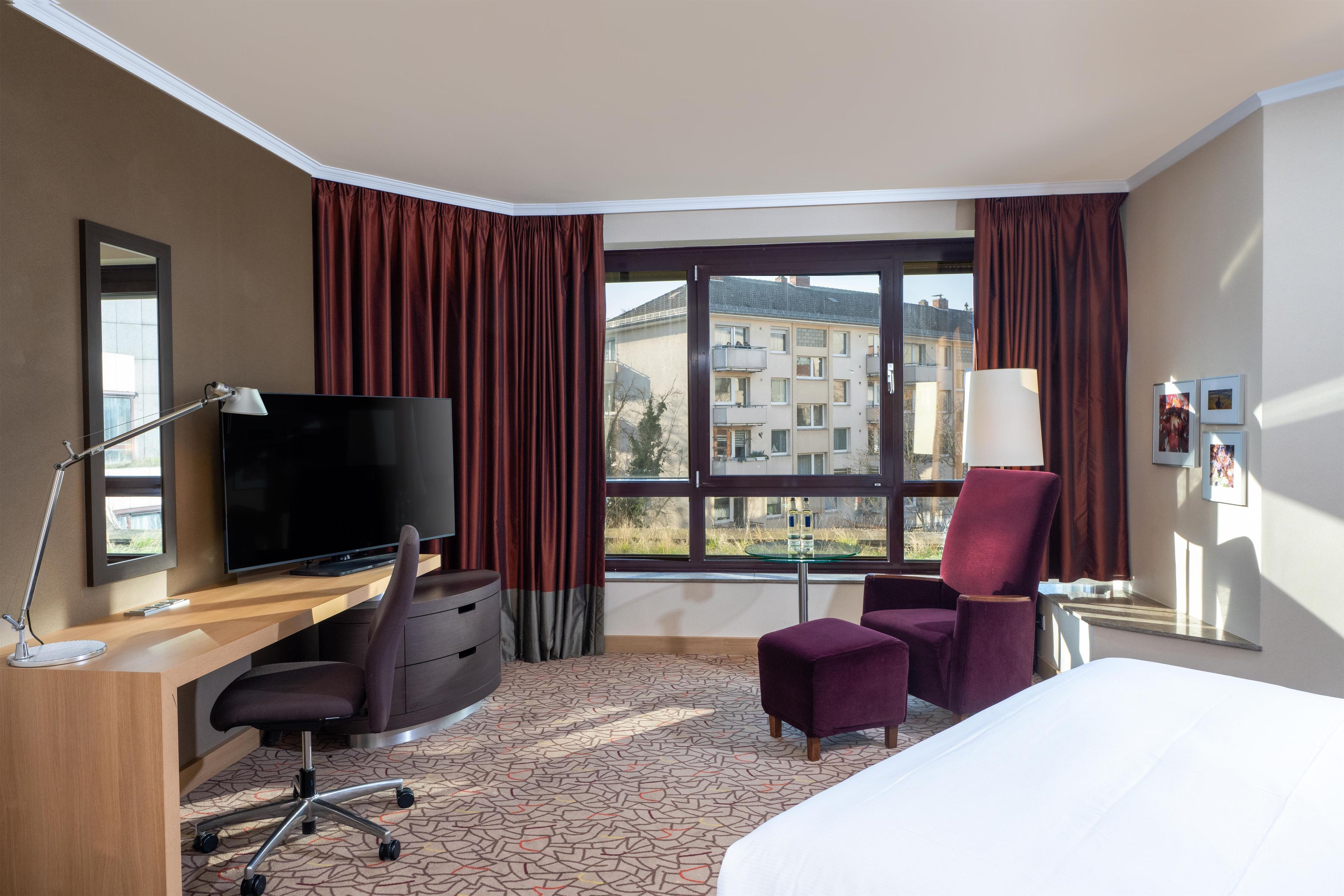 Una habitación de hotel con cama, escritorio, silla, televisión y ventana grande con cortinas.