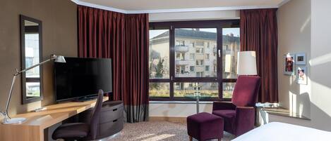 Una habitación de hotel con cama, escritorio, silla, televisión y ventana grande con cortinas.