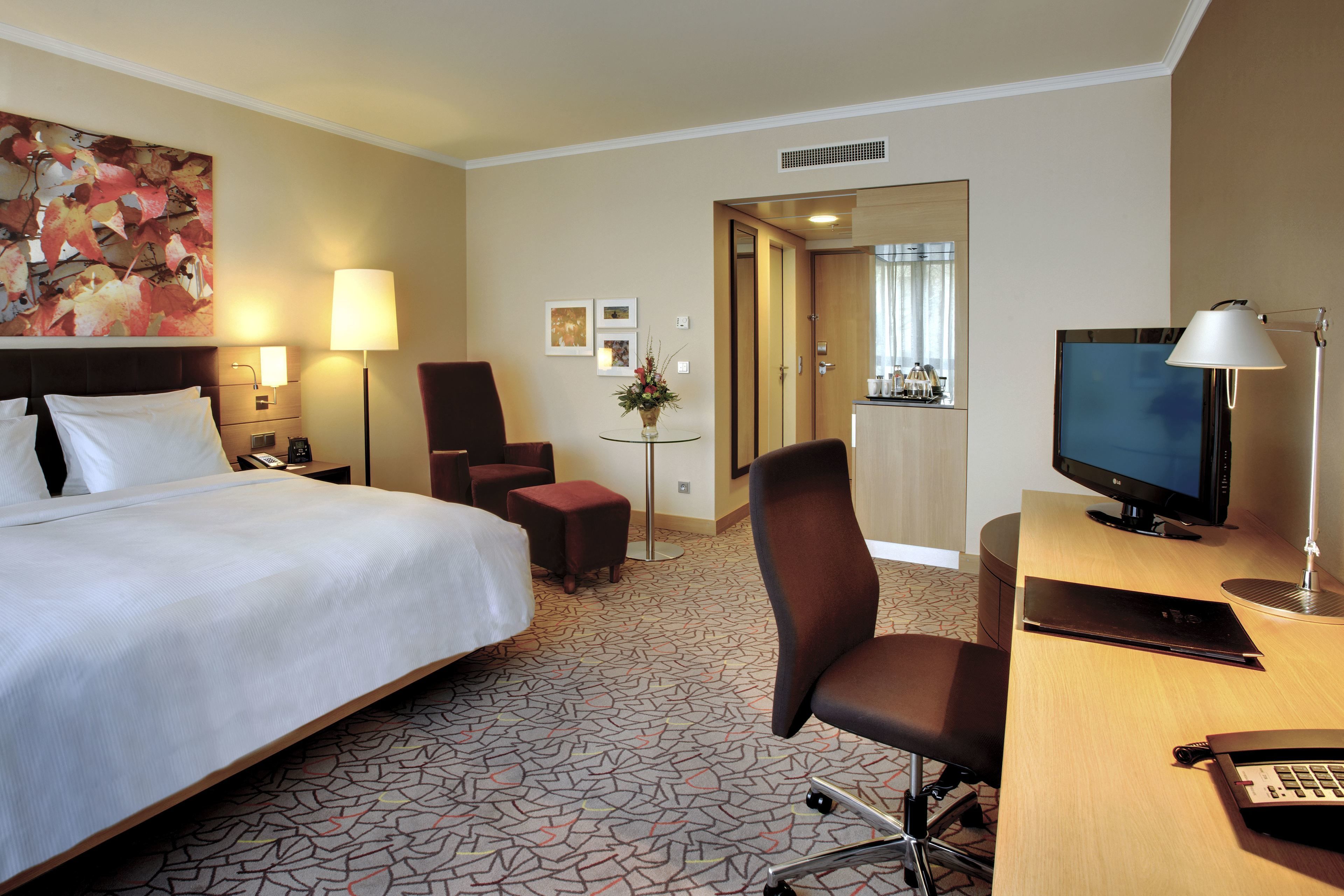 A hotel room with a large bed, a desk with a computer, a TV, and a chair. There is a small table with a vase of flowers.