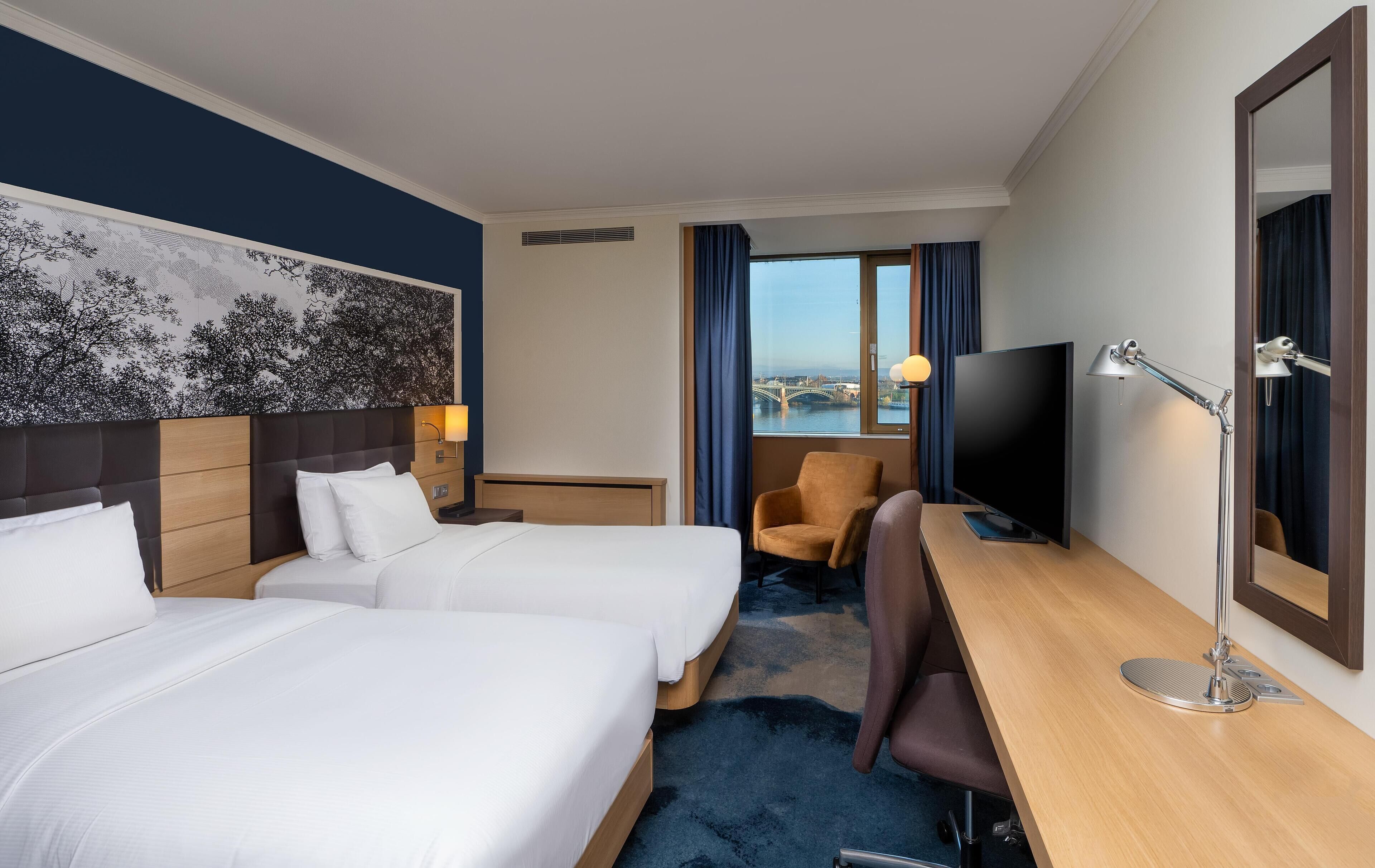 A hotel room with two beds, a desk, a chair, a TV, and a view of a bridge.