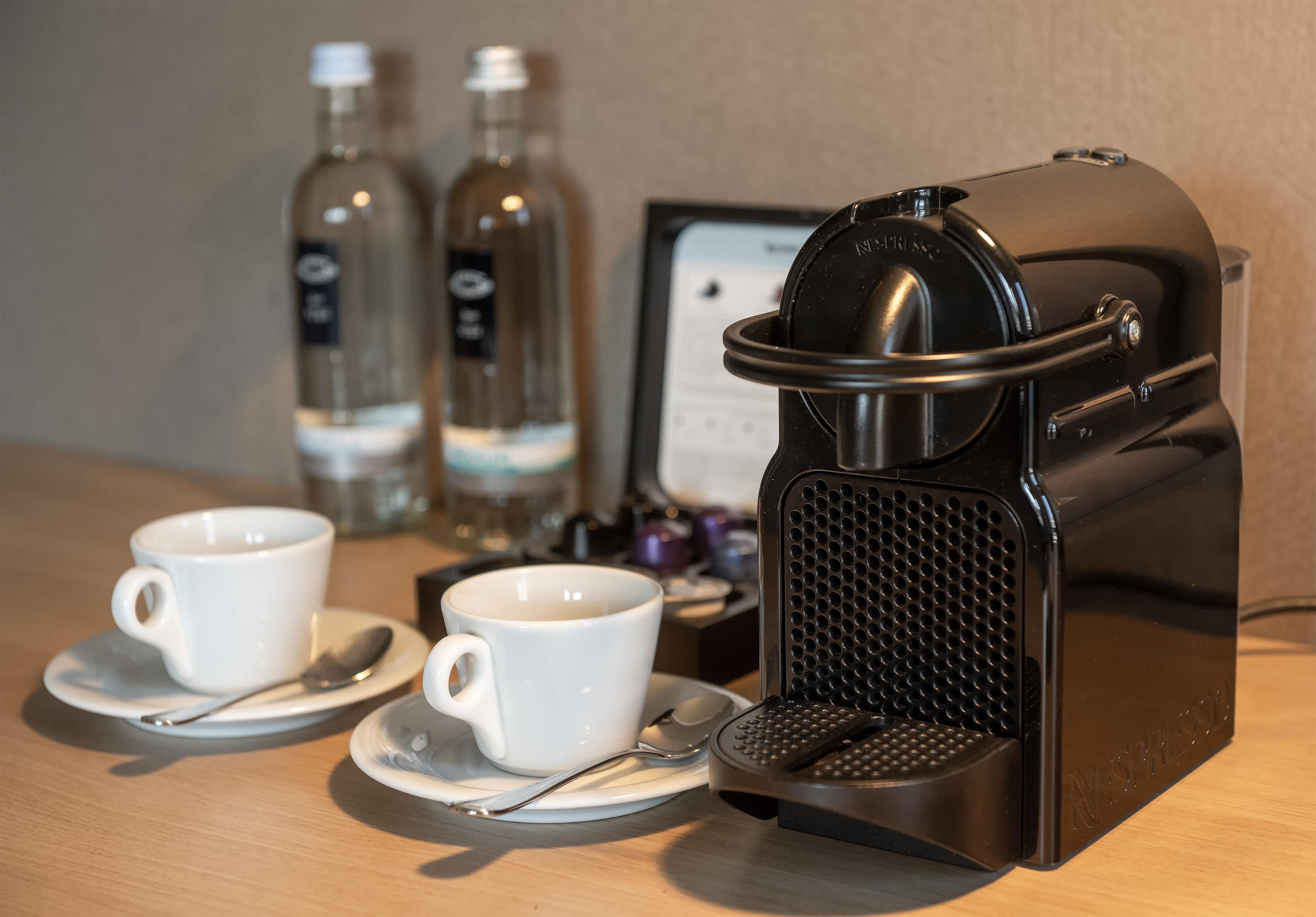 A coffee machine with two cups and spoons on a table, with bottles in the background.
