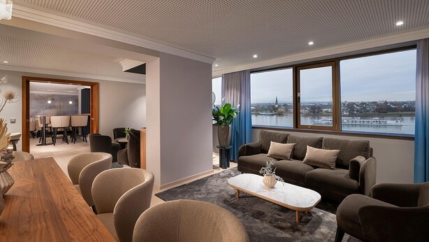 A modern living room with a large window, a dining area, and a view of a river. - Hilton Mainz (Mainz)
