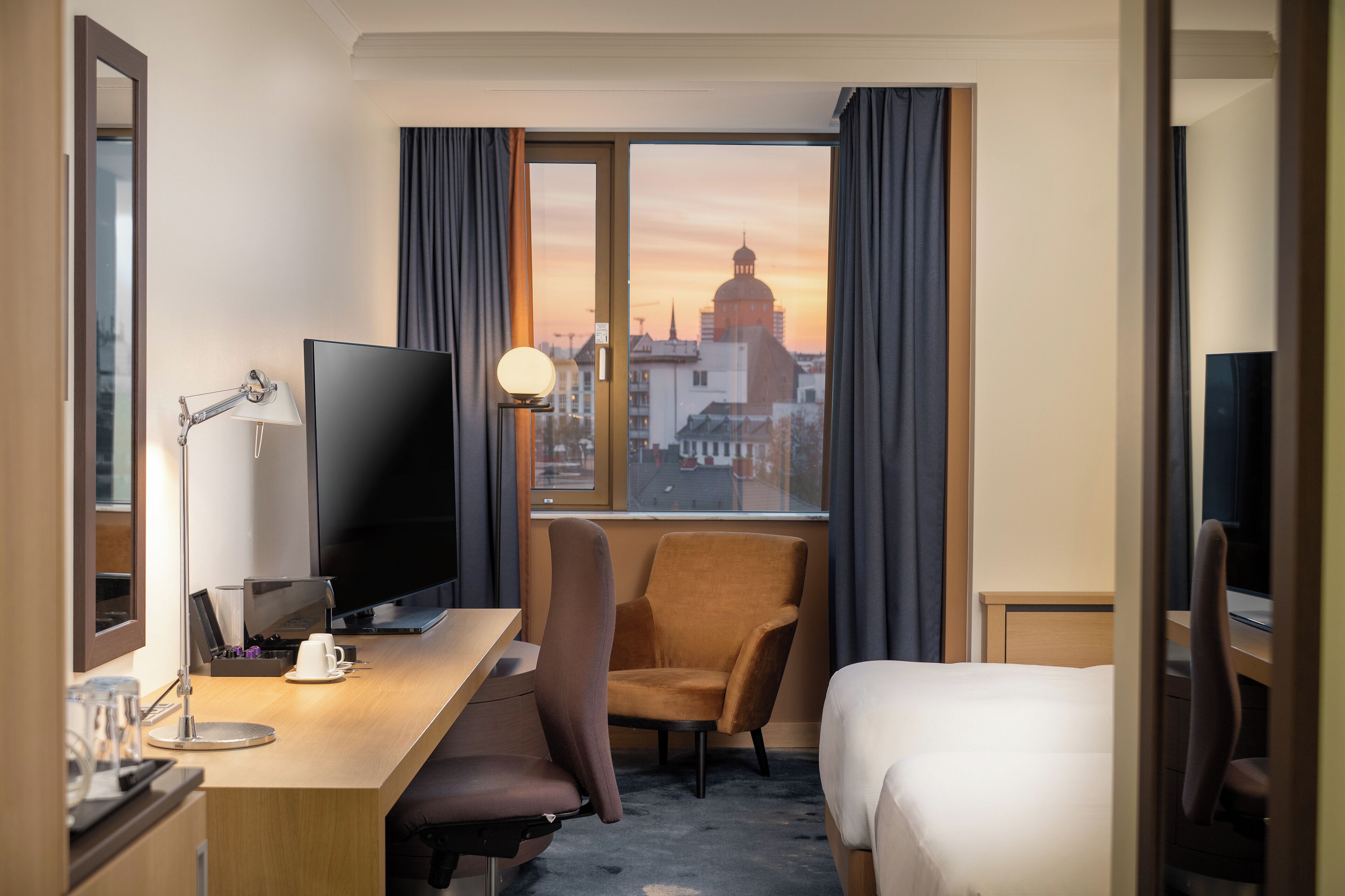 Habitación de hotel con televisor grande, escritorio con lámpara, una silla, cama y vista a un paisaje urbano al atardecer.