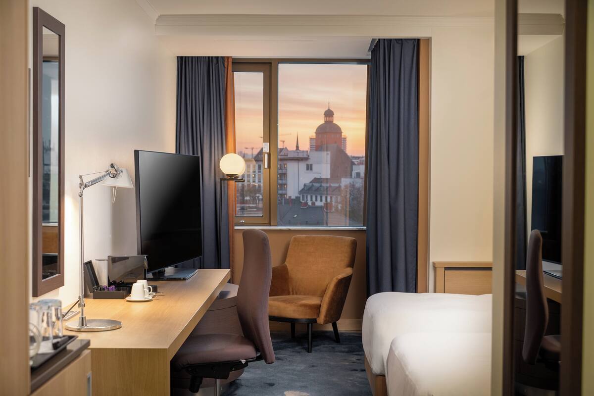 Habitación de hotel con televisor grande, escritorio con lámpara, una silla, cama y vista a un paisaje urbano al atardecer.