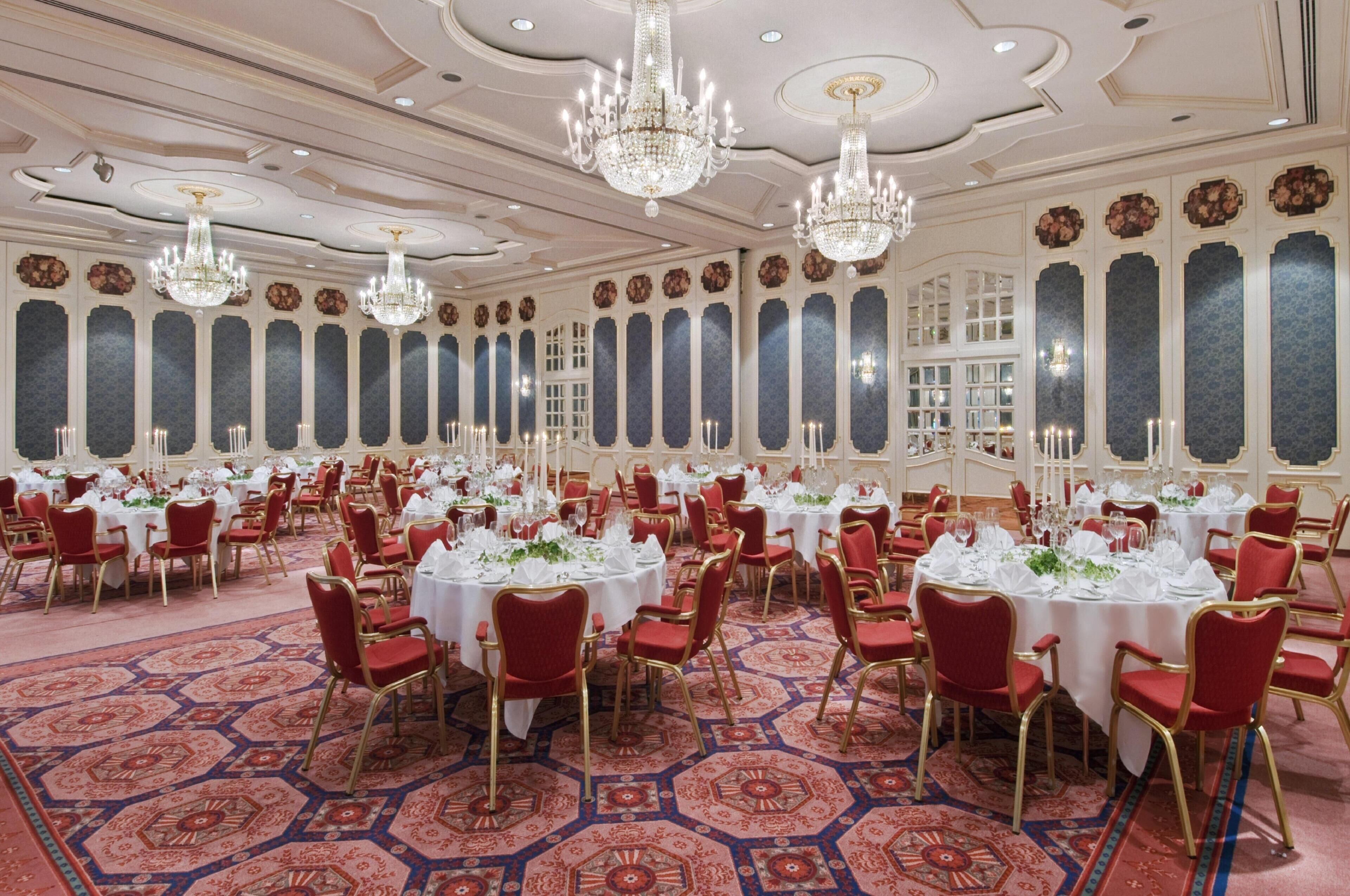 A banquet hall with round tables, red chairs, chandeliers, and patterned carpet.