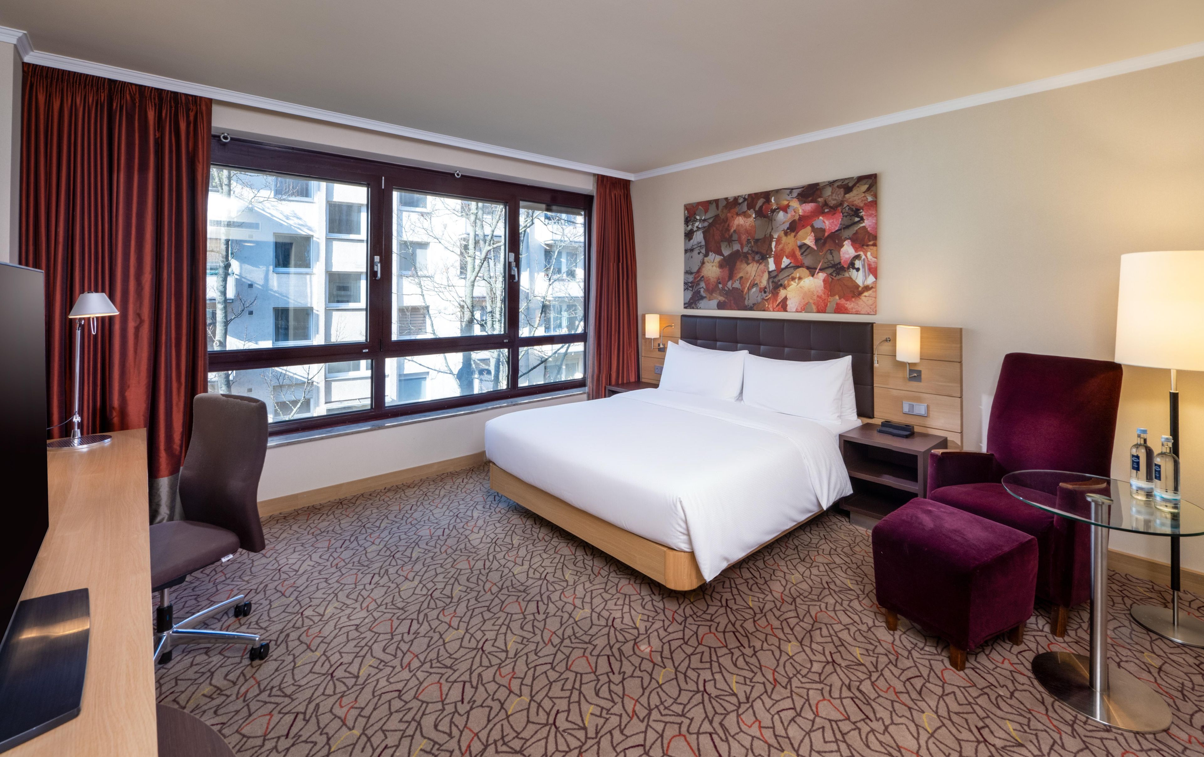 A hotel room with a large bed, a desk, a chair, a glass table, and a painting on the wall.