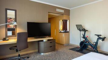 Habitación de hotel con cama, escritorio, silla, televisor, bicicleta estática y espejo.