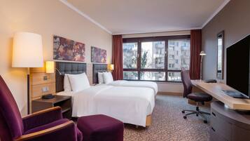 A hotel room with two beds, a desk, a chair, a TV, and artwork on the walls.