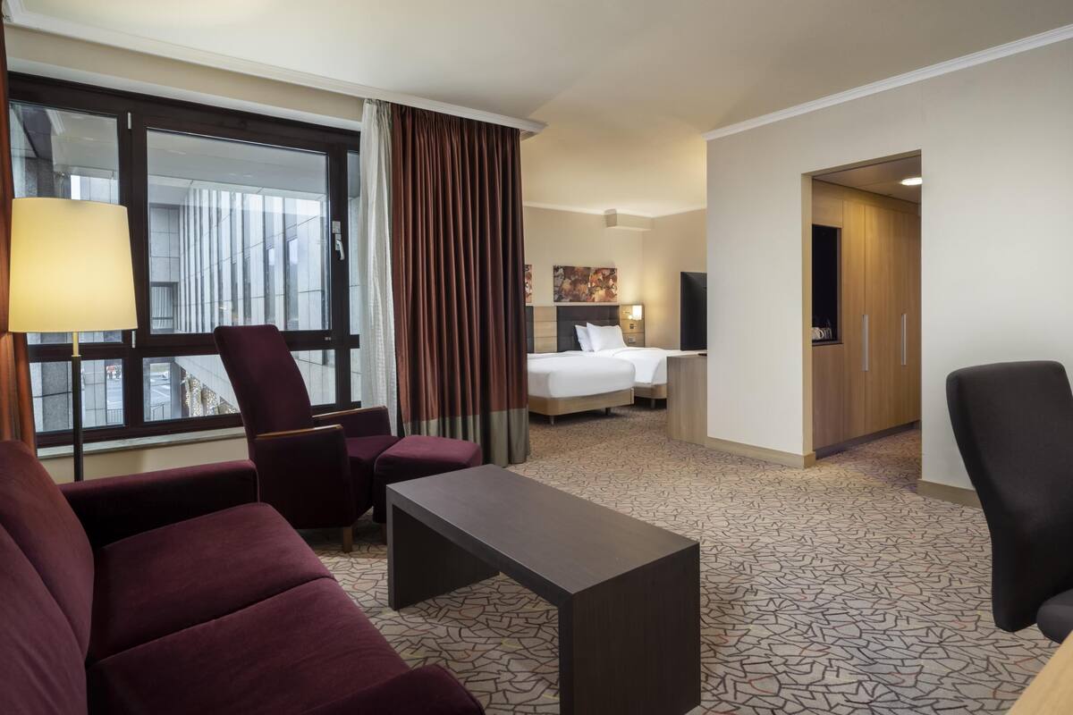 Habitación de hotel con cama, escritorio, dos sillones, televisor y ventana con cortinas.