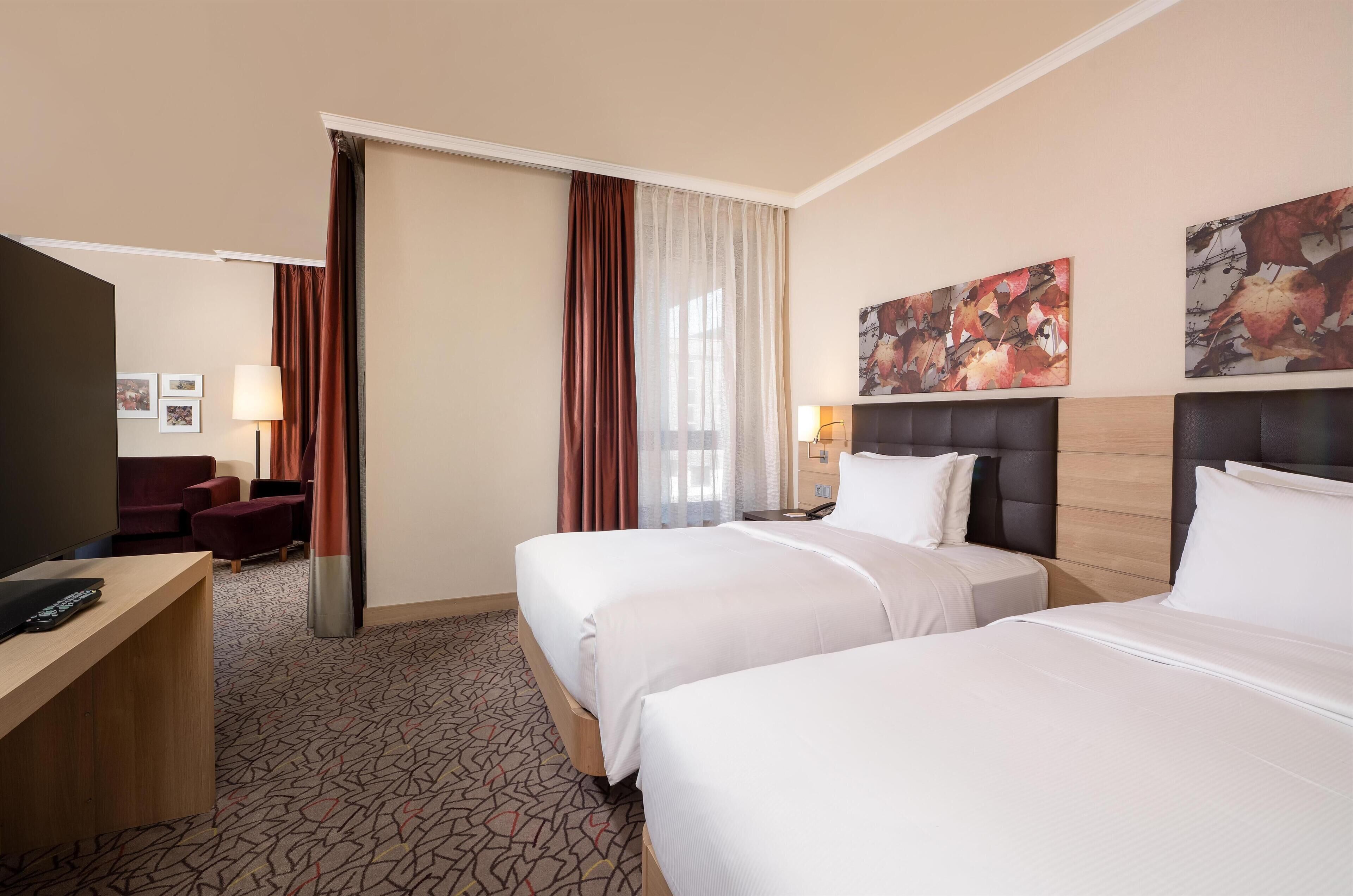 Habitación de hotel con dos camas, un escritorio, un televisor y una zona de estar con sofá y sillones.