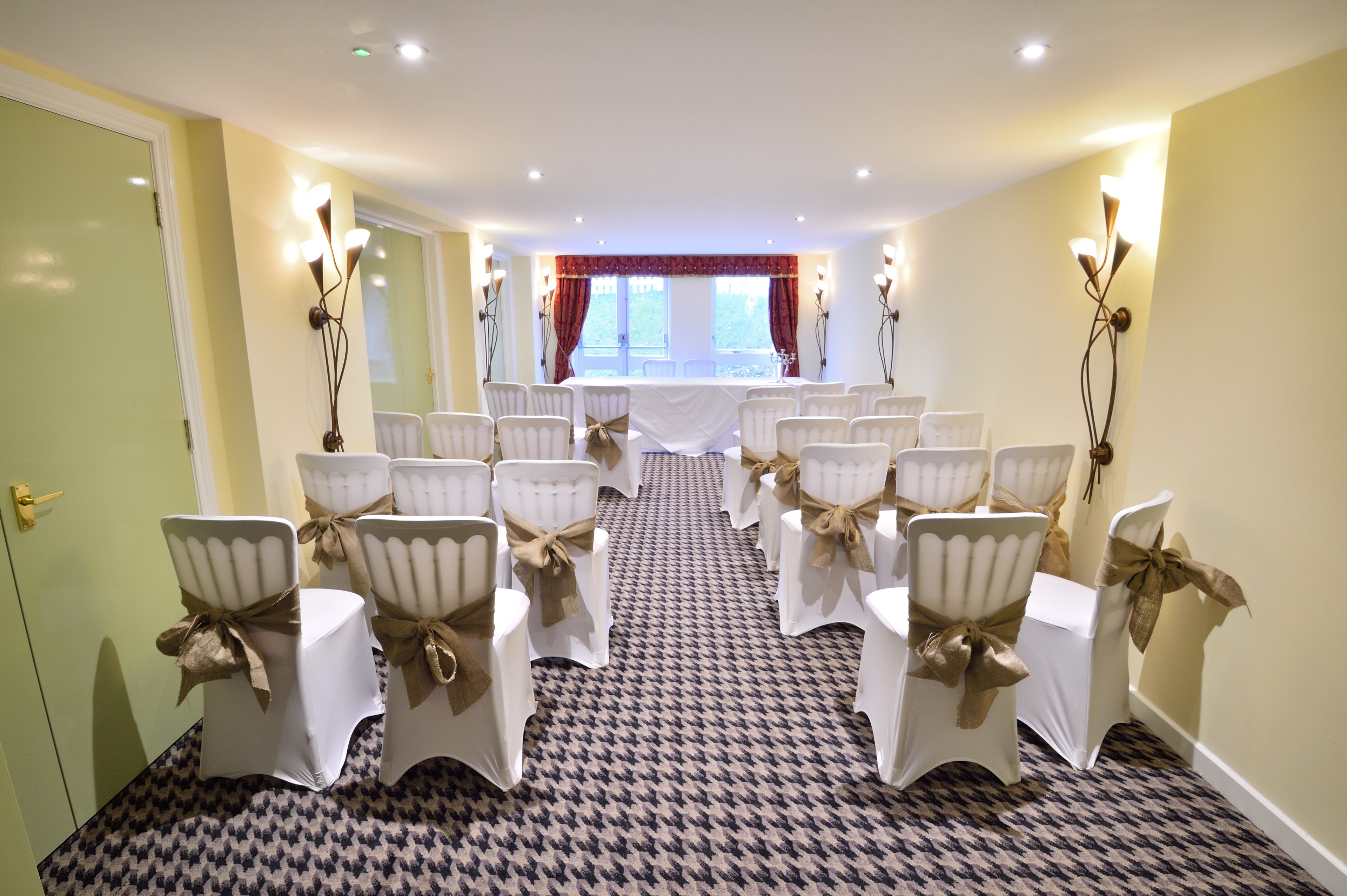 A conference room with rows of white chairs tied with brown ribbons, a patterned carpet, and a view of a window with red curtains.