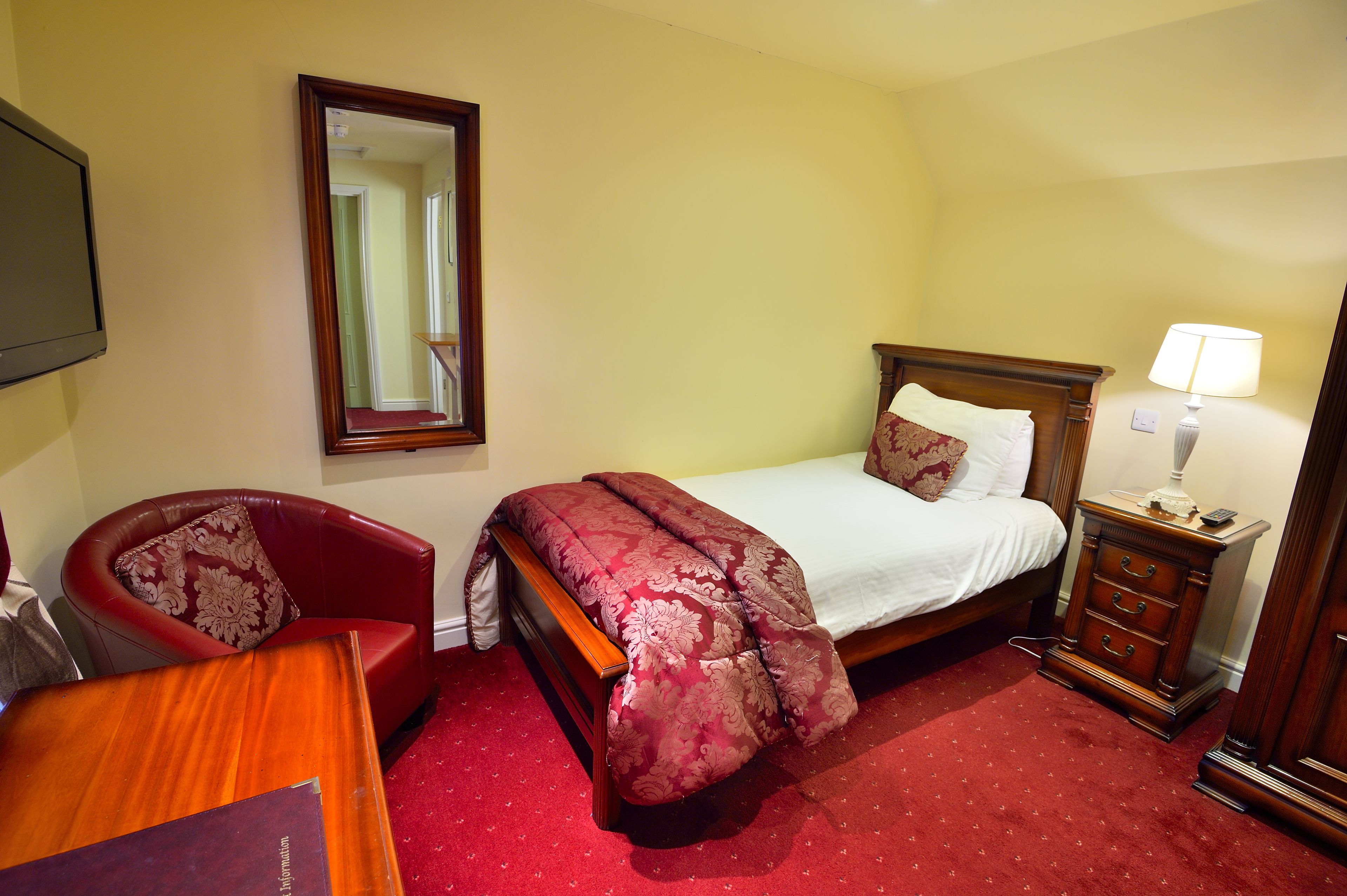 Una habitación de hotel con una cama, una mesita de noche de madera con una lámpara, un sillón rojo y un espejo grande en la pared.