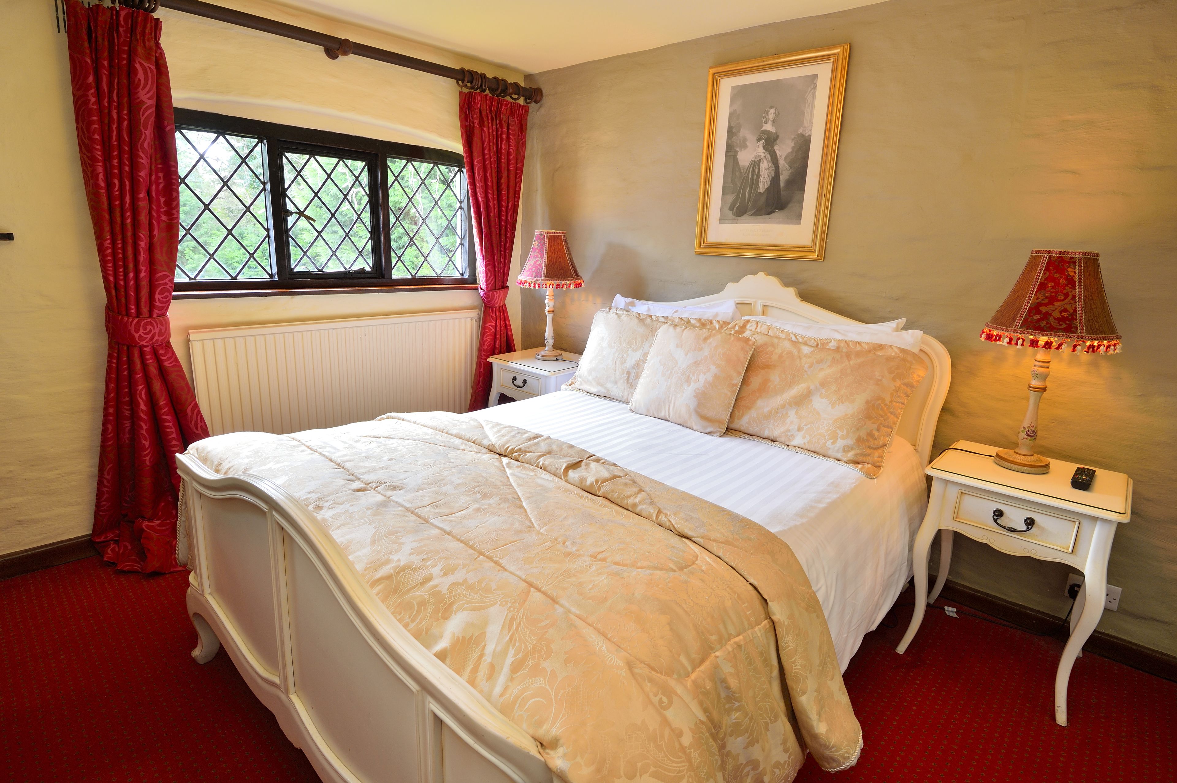 A neatly made bed with a white and beige bedspread, two bedside tables with lamps, a radiator, and a window with red curtains.