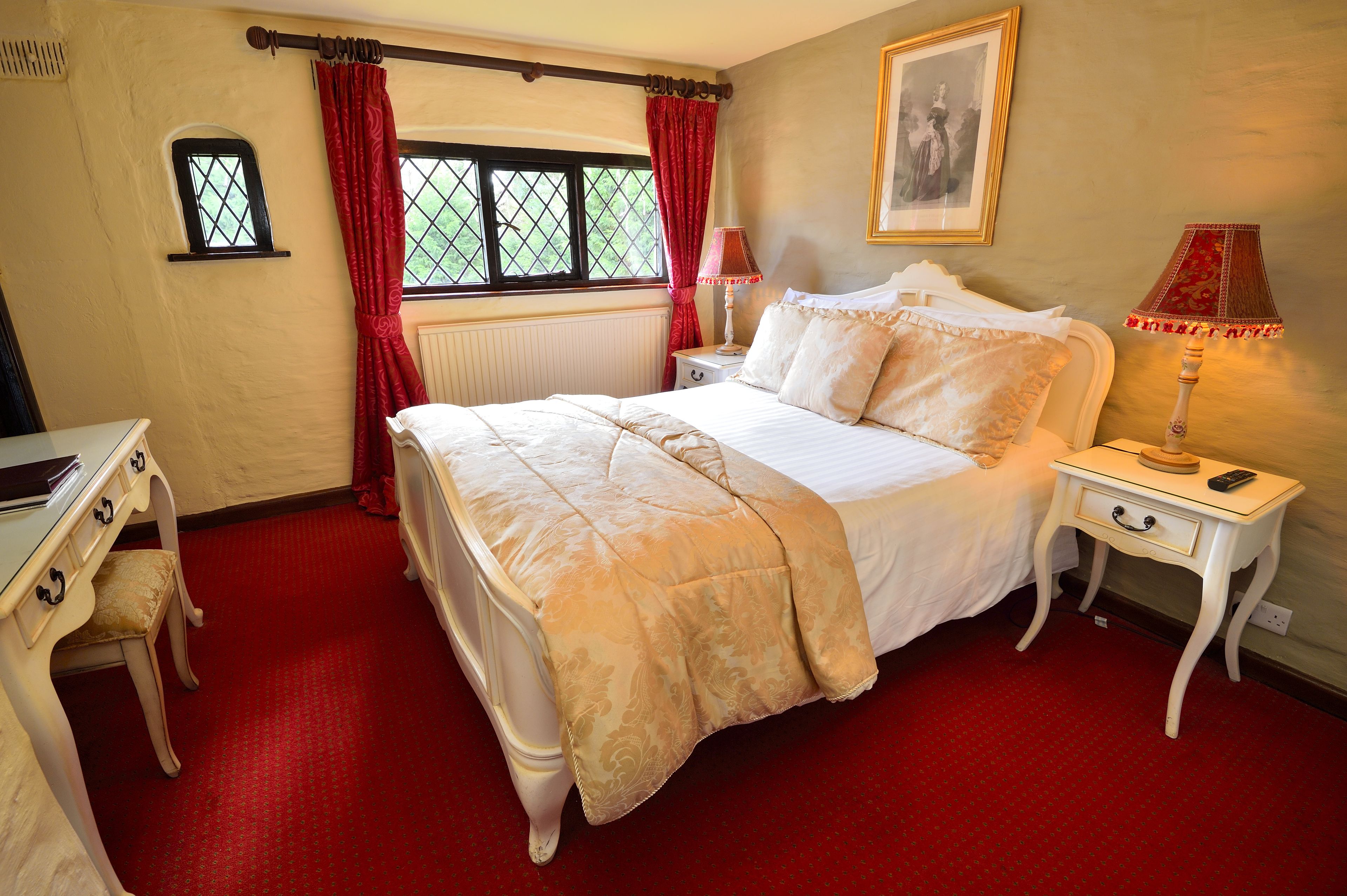 A bedroom with a large bed, a bedside table with a lamp, a small dresser, and a window with red curtains.