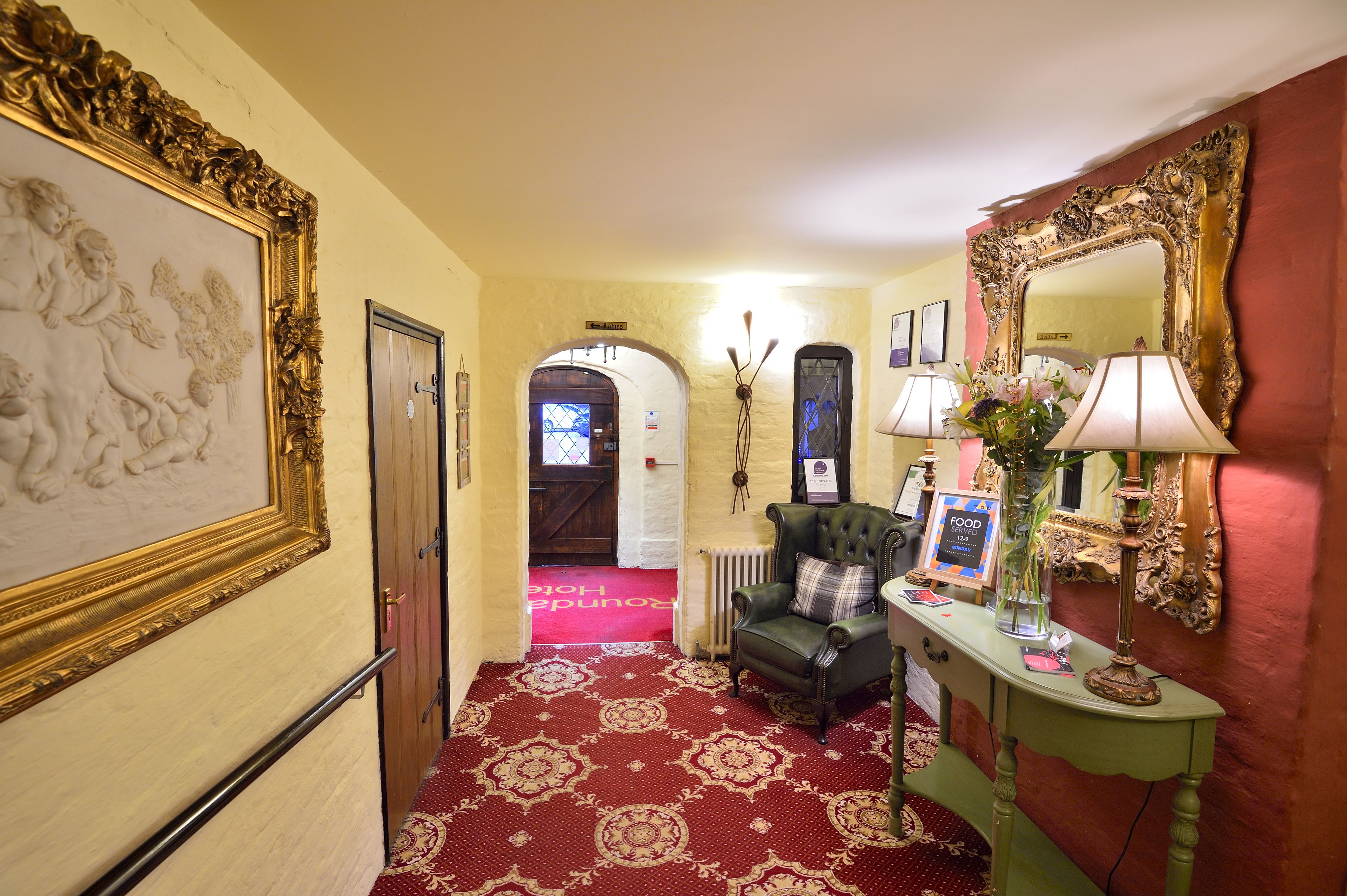 A hallway with a red carpet, a green armchair, a table with a lamp, a mirror, and a framed picture on the wall.