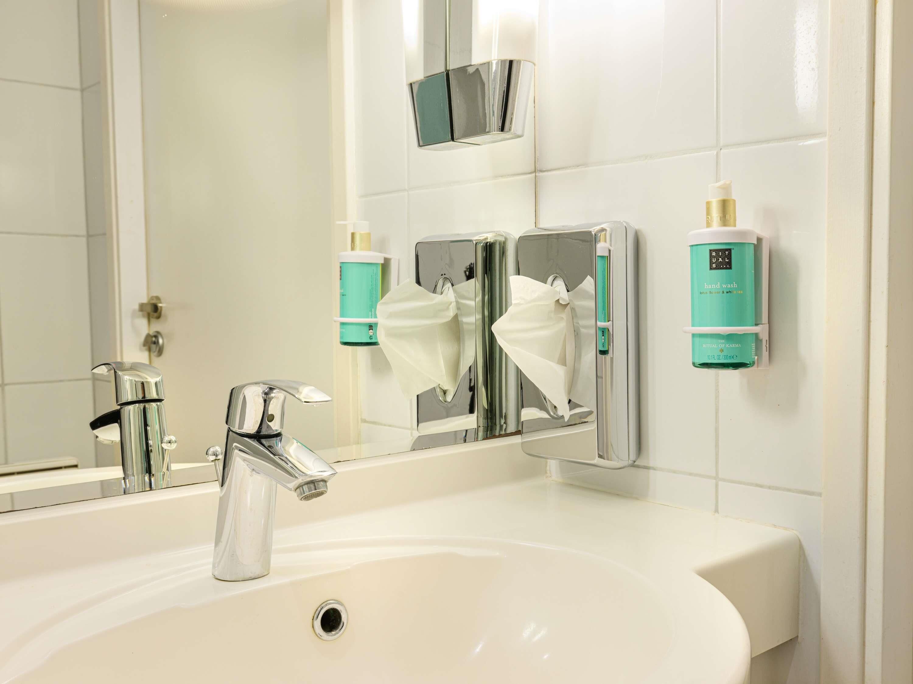 Un baño con lavamanos blanco, grifo cromado y espejo. Hay dos botellas de jabón en la pared.