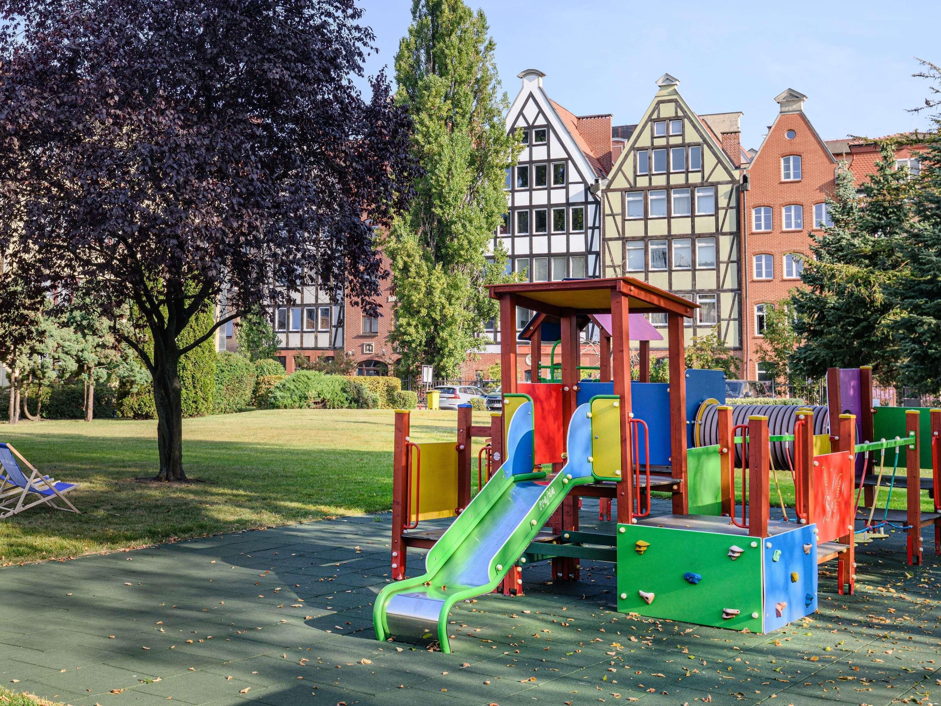 A playground with slides and climbing structures, surrounded by a park with grass and trees, and buildings in the background.