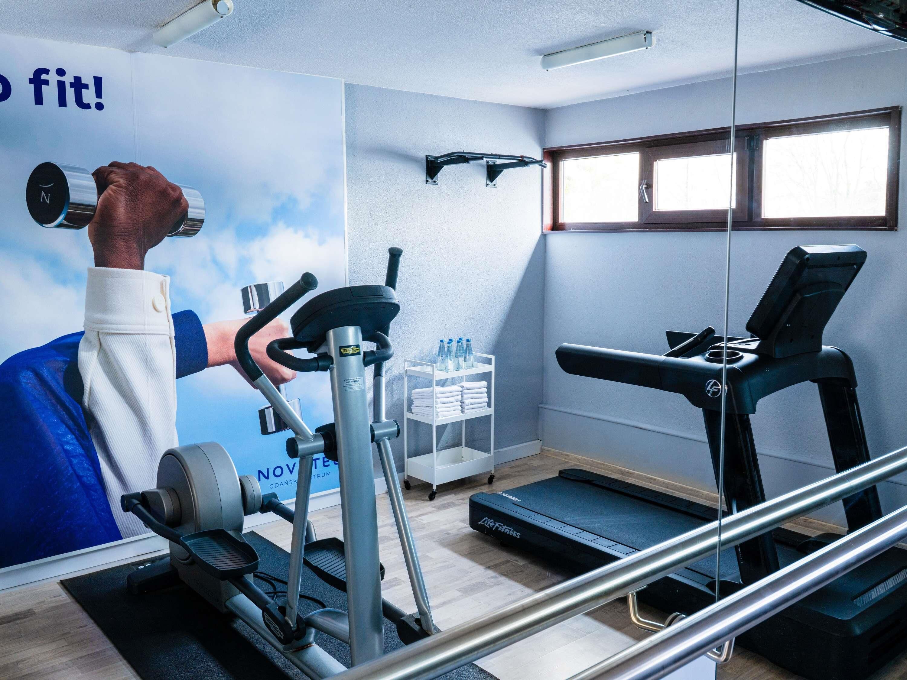Un gimnasio con cinta de correr, elíptica y pesas. Hay un cartel de una persona levantando pesas, un dispensador de agua y una ventana con persianas.