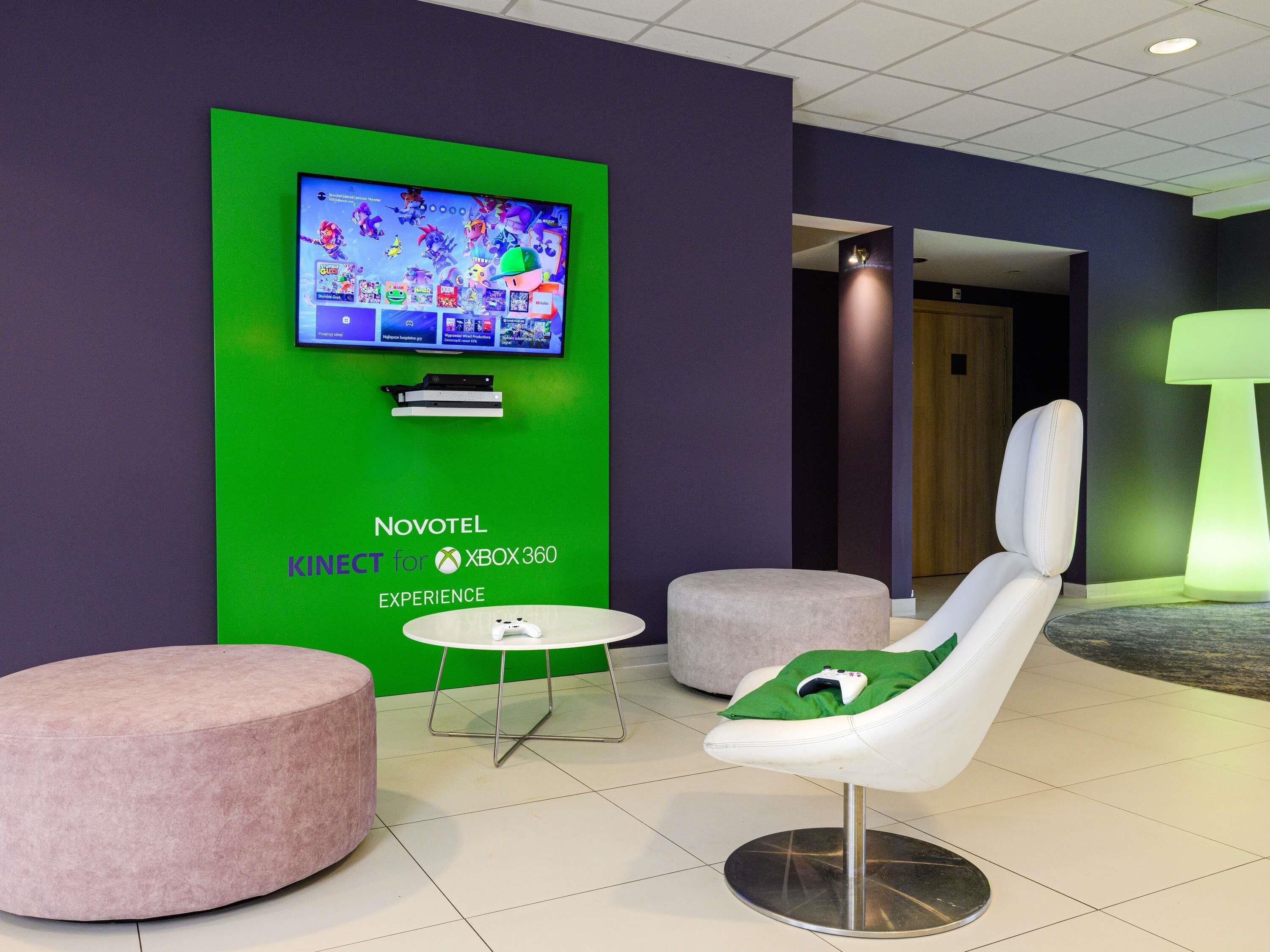A hotel lobby with a green wall displaying a Novotel Xbox 360 experience sign, a white gaming chair with a game controller, and two round, pink ottomans.