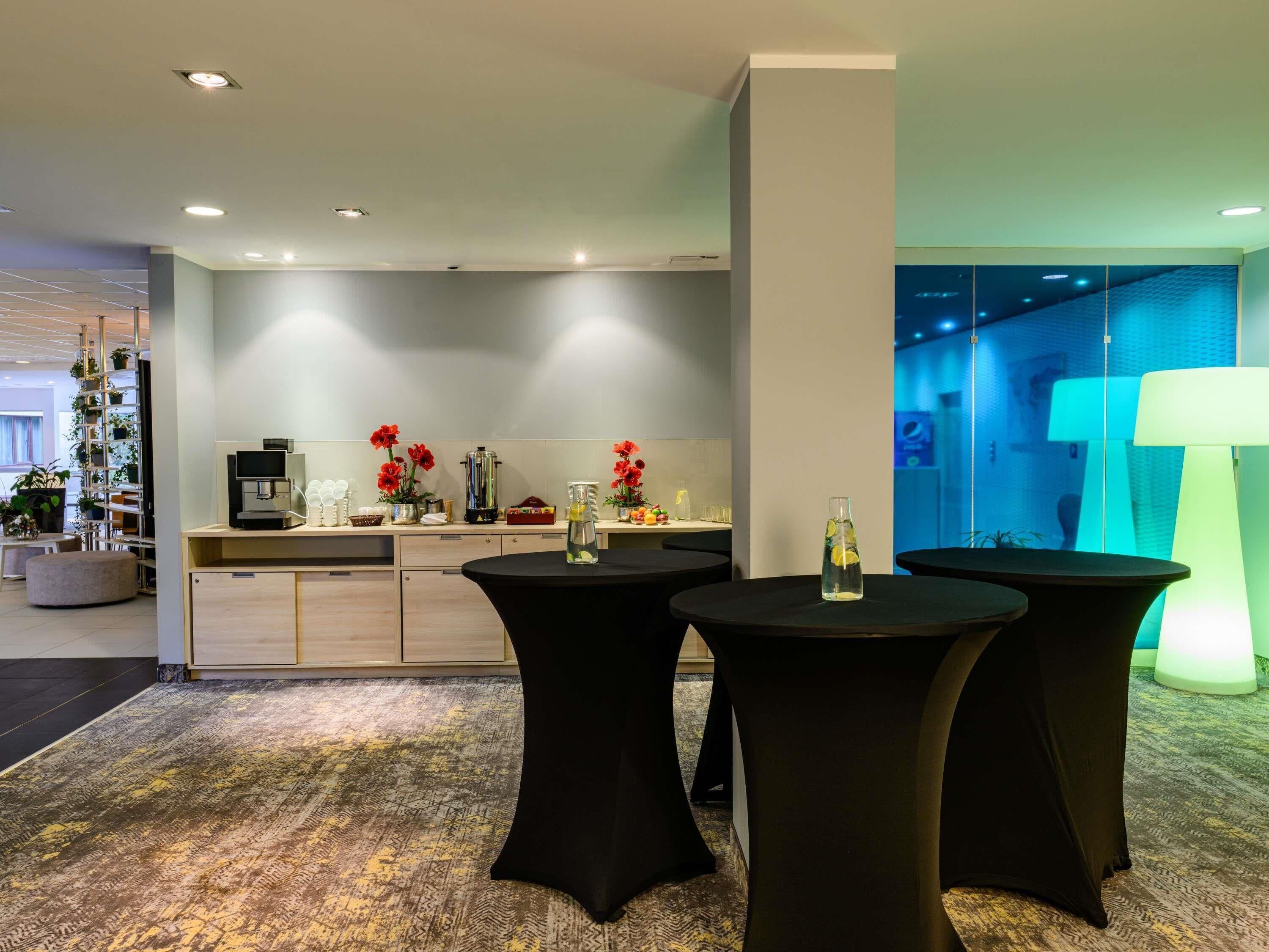 A modern hotel lobby with a coffee station, seating area, and a view into a room with a blue-lit wall.
