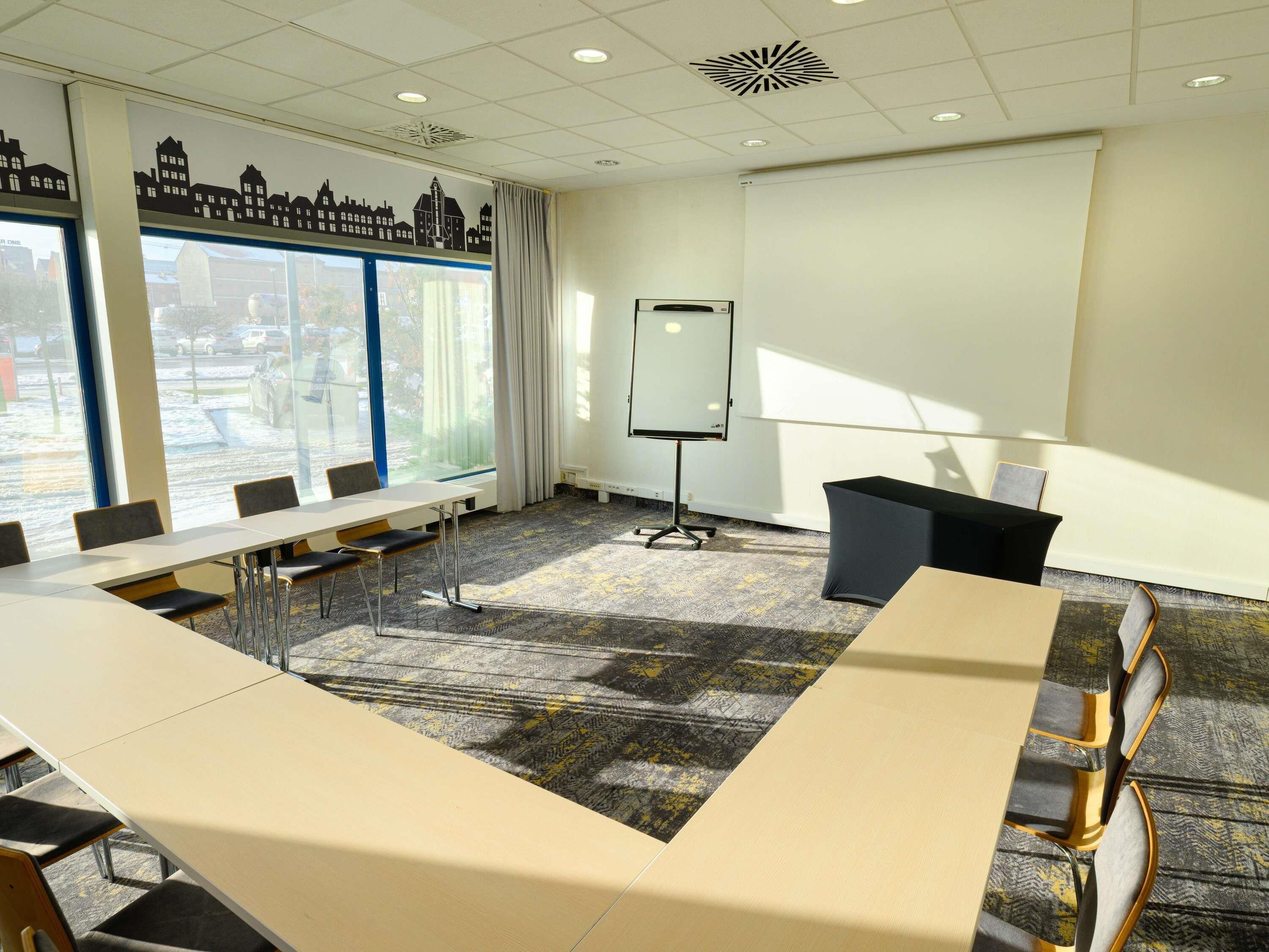 A conference room with a long table, chairs, a projector screen, and large windows with a view of buildings.