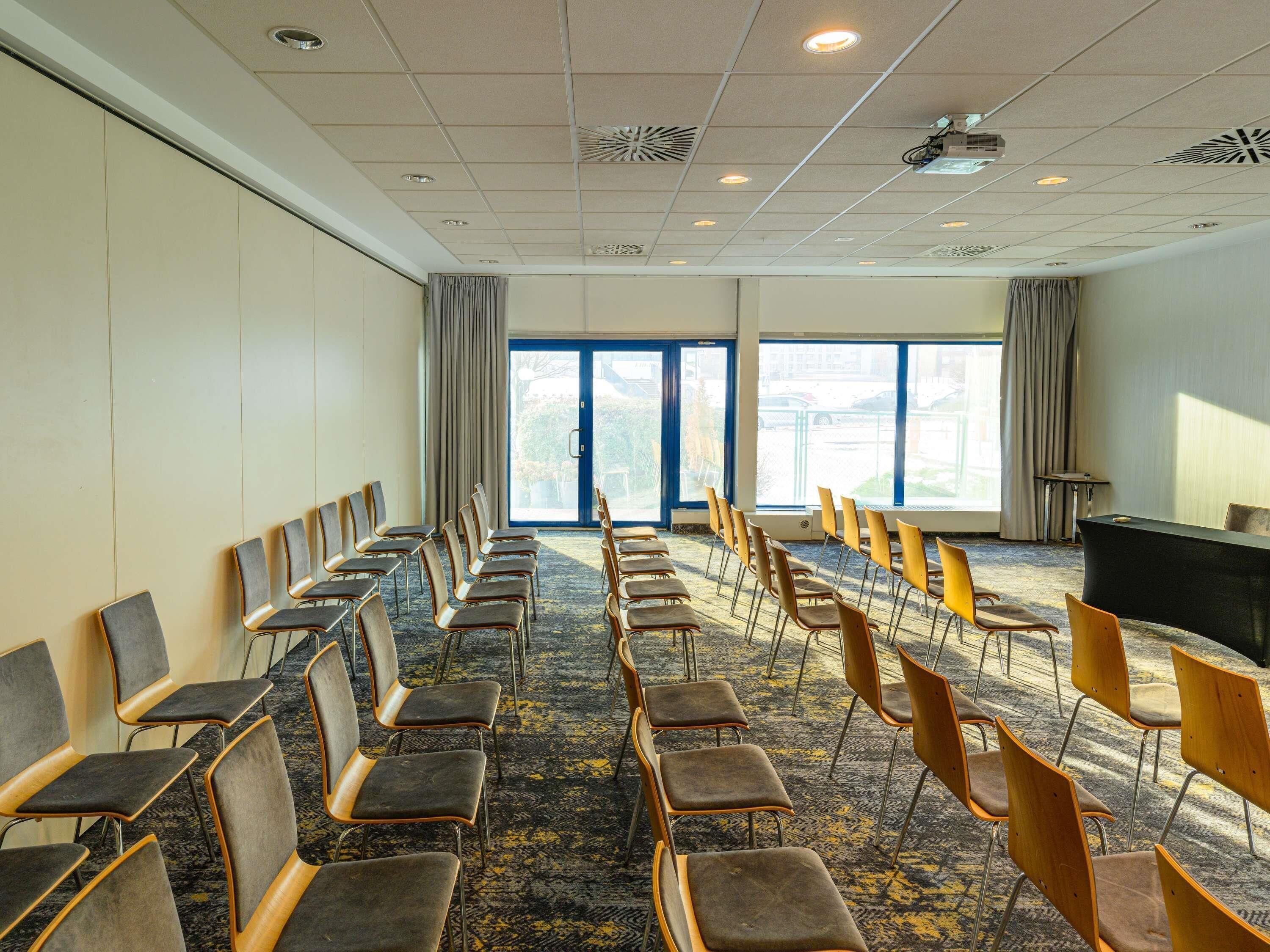 Une salle de conférence avec des rangées de chaises, un projecteur et de grandes fenêtres.