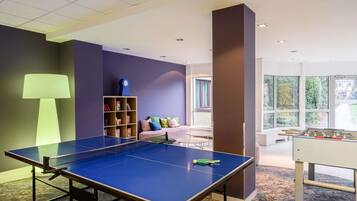 A ping pong table, a bookshelf, a sofa, and a foosball table in a room with large windows.