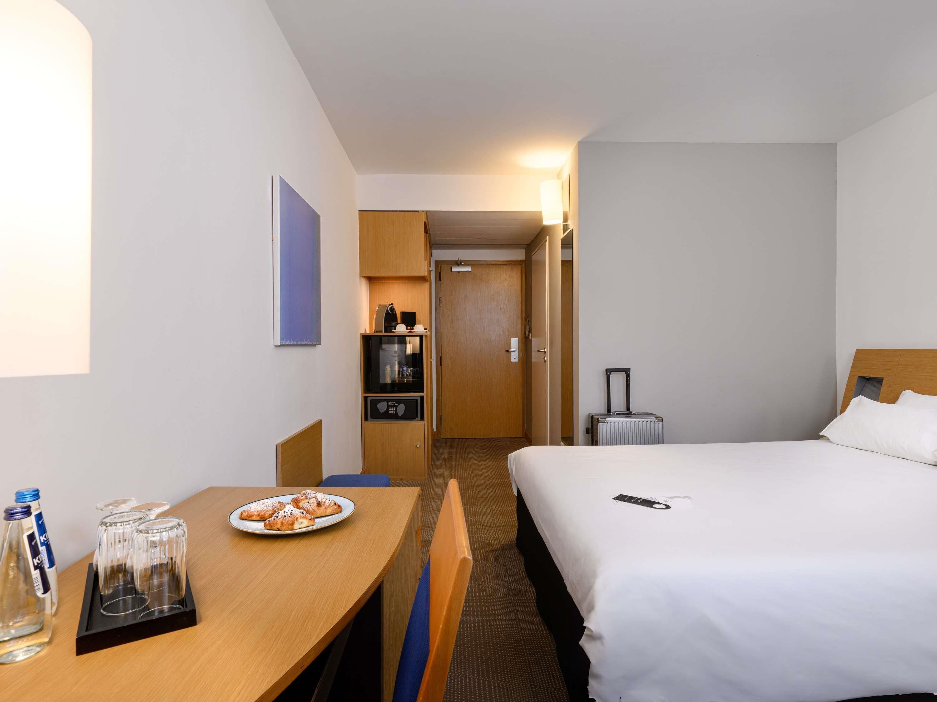 A hotel room with a bed, a desk with a plate of pastries, a coffee machine, and a suitcase.
