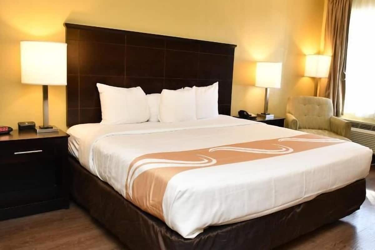 A hotel room with a large bed, bedside lamps, a chair, and an air conditioning unit.