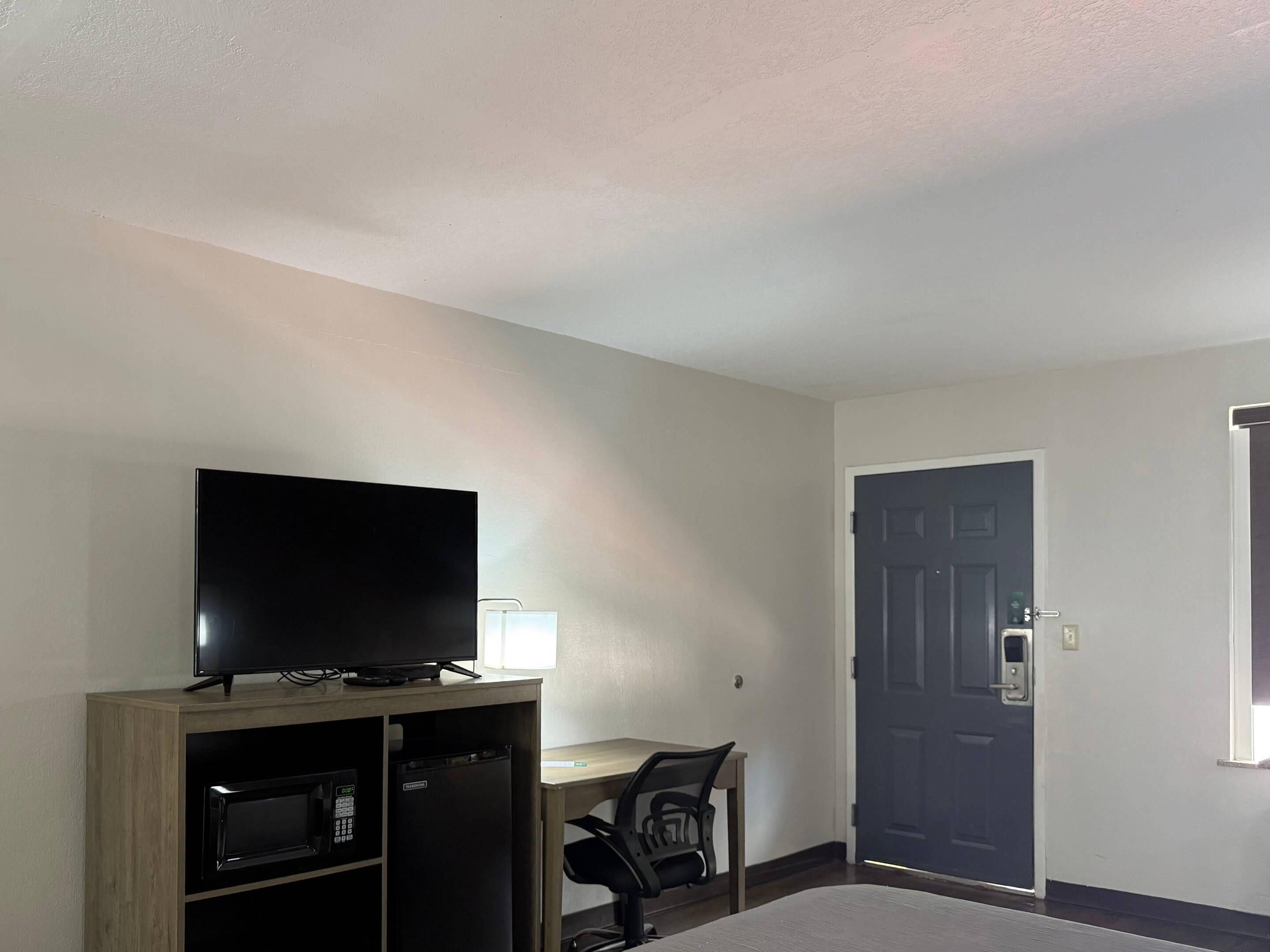 A room with a flat-screen TV on a wooden cabinet, a desk with a chair, and a door.