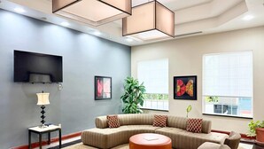 A modern living room with a sectional sofa, a round ottoman, a flat-screen TV, and large windows with blinds. - Quality Inn & Suites Greenville I-65 (Greenville)