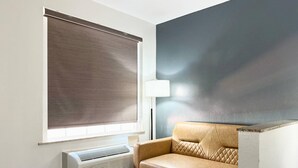 A modern room with a beige sofa, a large window with a blind, a floor lamp, and a wall-mounted air conditioner. - Quality Inn & Suites Greenville I-65 (Greenville)