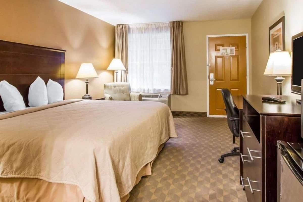 A hotel room with a large bed, two bedside tables with lamps, a chair, a desk with a chair, a door, and a television.