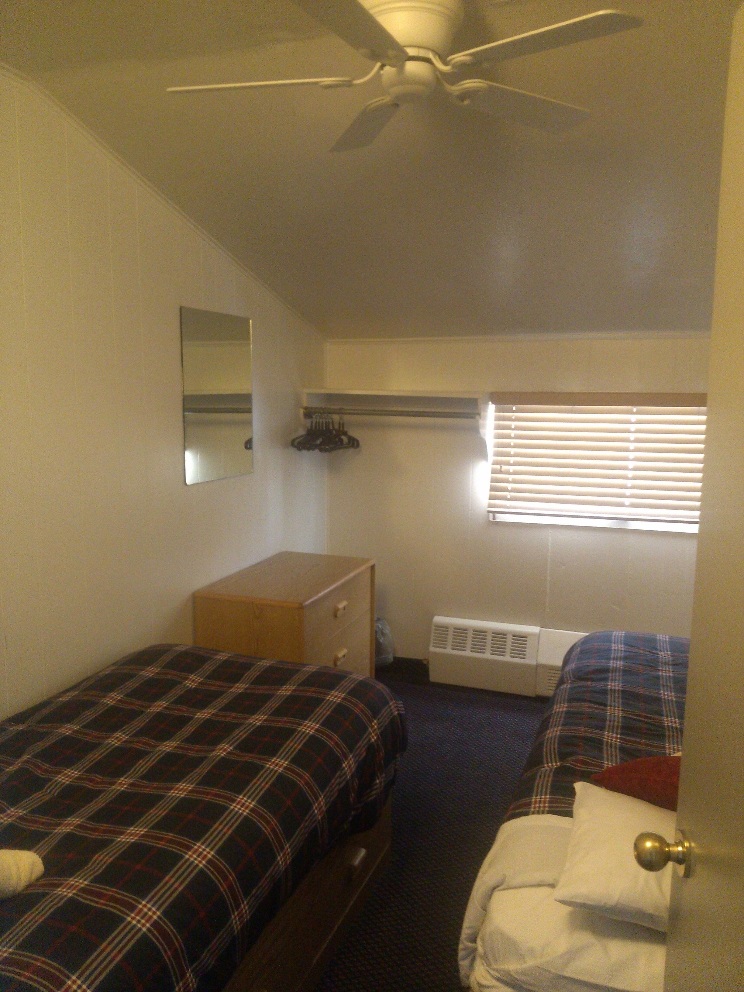 A small, compact room with two single beds, a ceiling fan, a radiator, and a window with blinds.