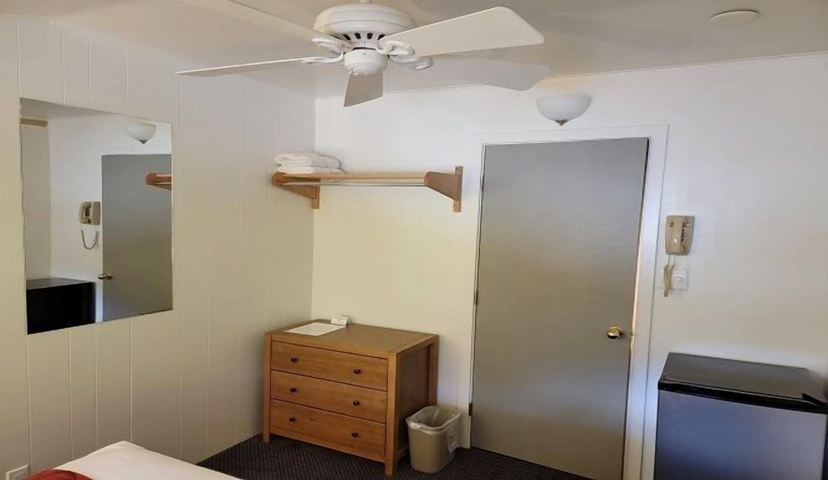 A small, clean room with a wooden dresser, a mirror, a ceiling fan, and a door.