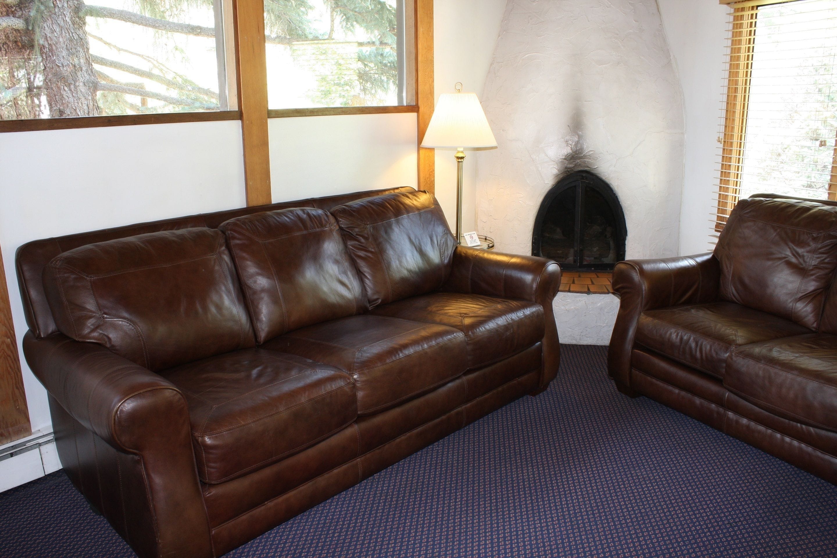Una sala de estar con un sofá de cuero marrón, un sillón a juego, una chimenea y una ventana con persianas.