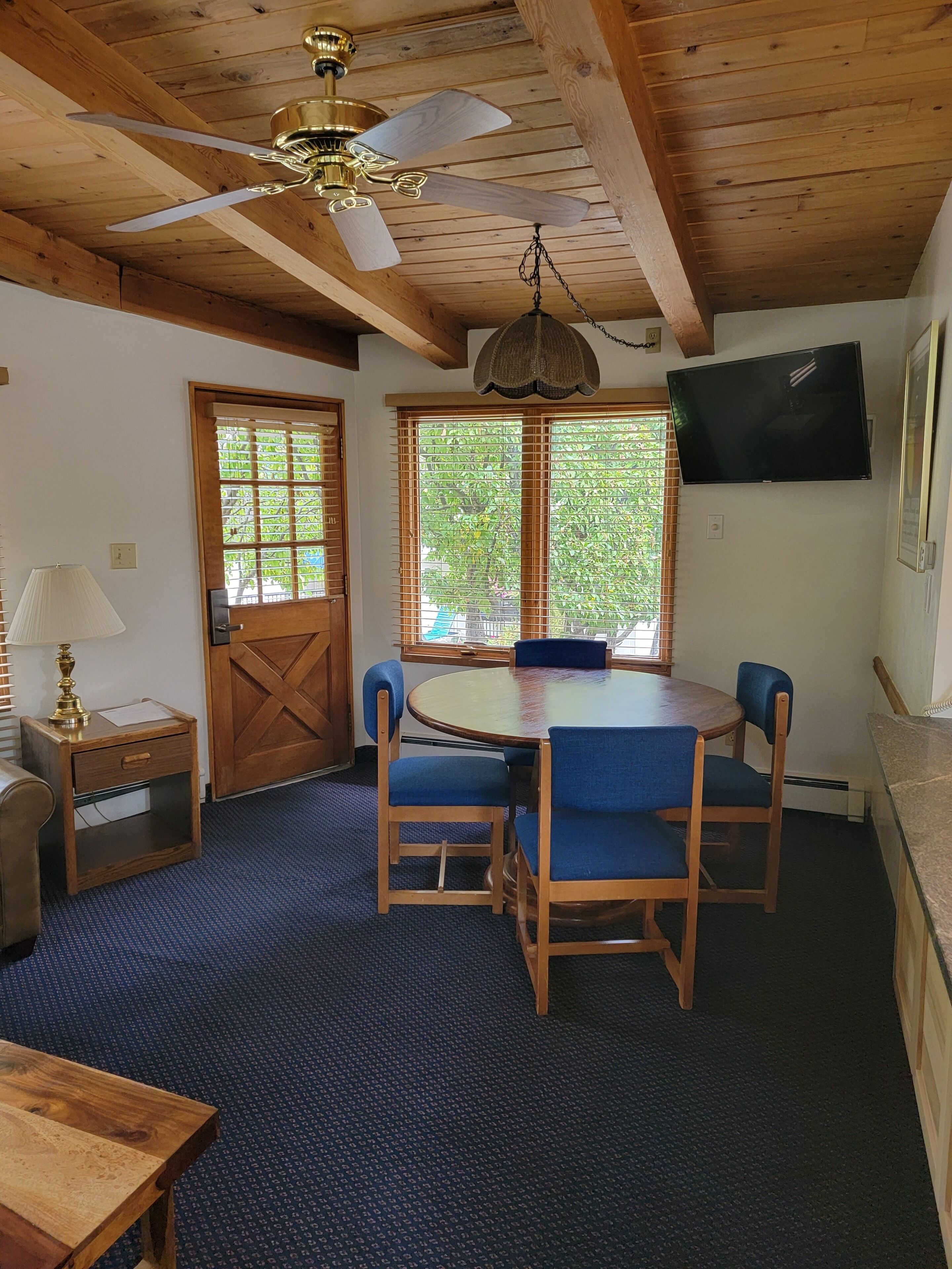 Ett rum med ett träpanelat tak, en takfläkt, en tv som är monterad på väggen, ett runt bord med blå stolar och en dörr med ett korsformat träornament.