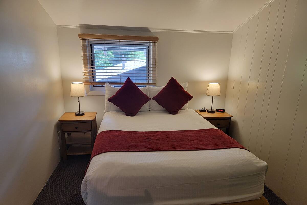Una cama bien hecha con una manta roja y dos almohadas, flanqueada por mesitas de noche con lámparas, en una habitación con paredes blancas y una ventana con persianas.