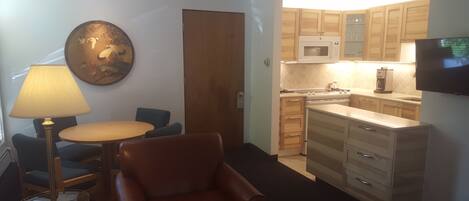 A compact living space with a kitchenette, a dining area with a table and chairs, and a seating area with a chair and a lamp.