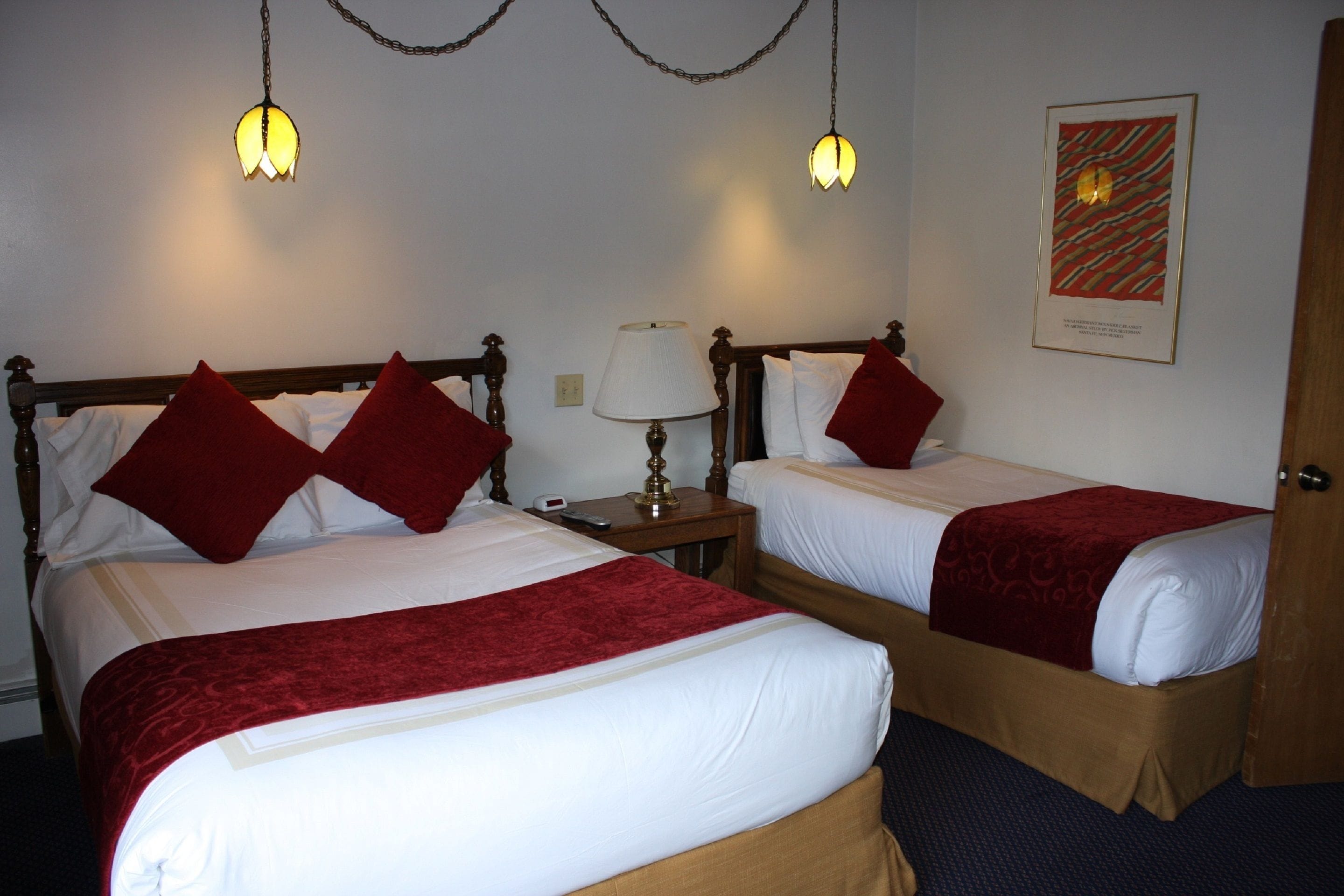Une chambre d’hôtel avec deux lits simples, chacun recouvert d’une couverture rouge et agrémenté d’oreillers. On y trouve une table de chevet avec une lampe, et un tableau encadré est accroché au mur.