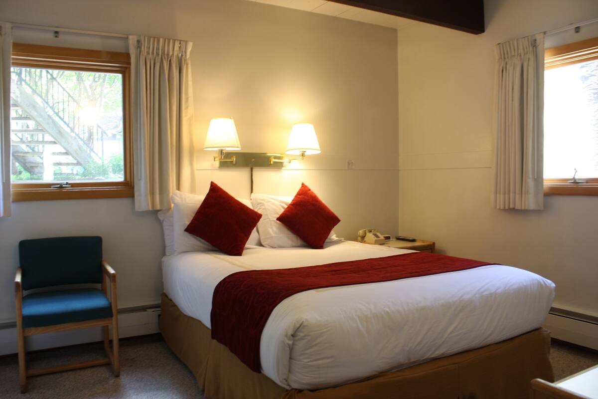 Una habitación de hotel con una cama, dos lámparas de noche, una silla y una ventana con cortinas.