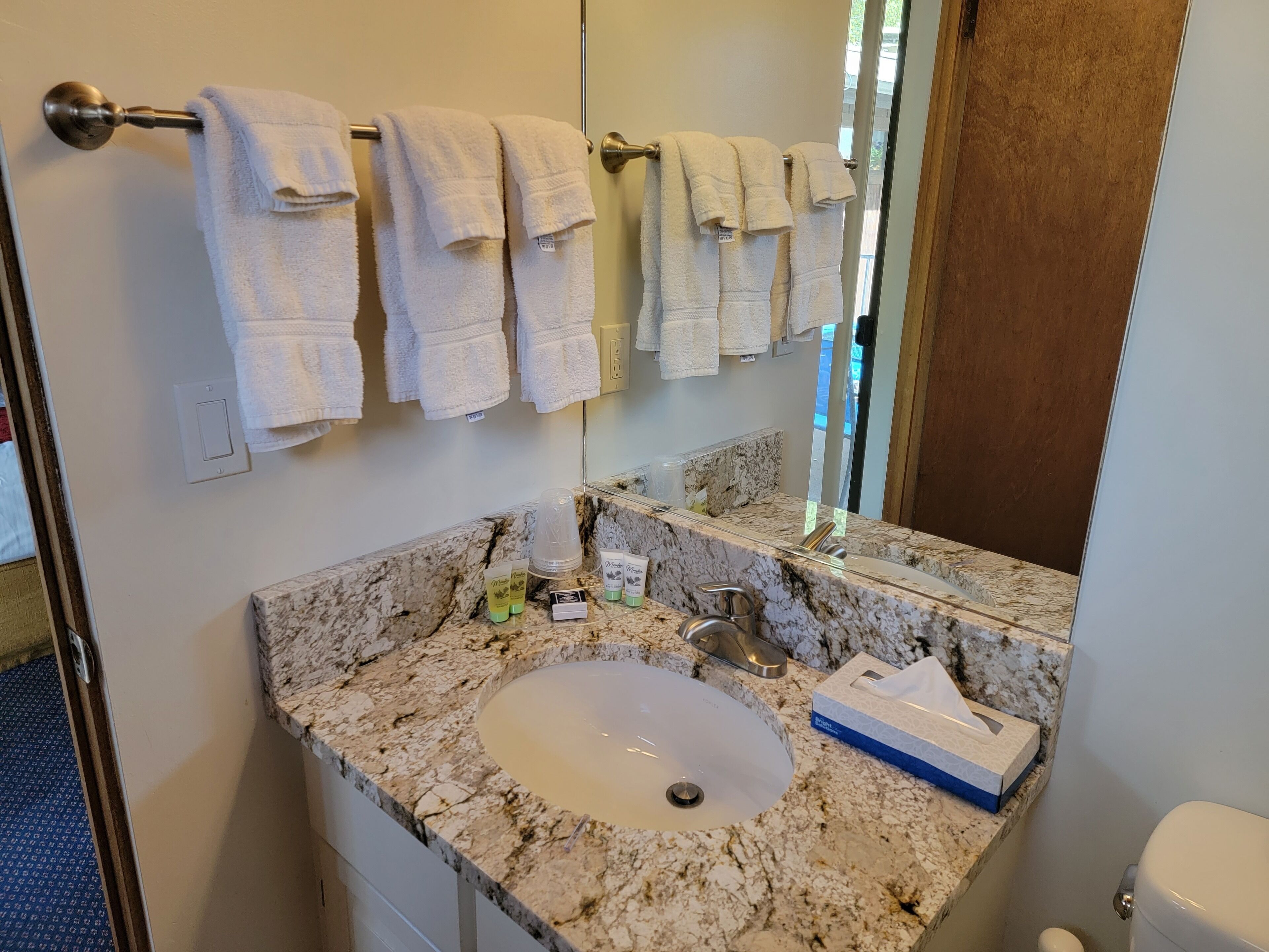 A bathroom with a marble sink, a mirror, a towel rack with multiple white towels, toiletries, and a toilet.