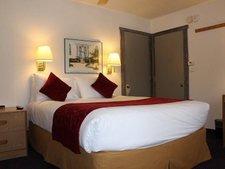 A neatly made bed with white linens and red pillows, two bedside lamps, a framed picture on the wall, and a closed door.