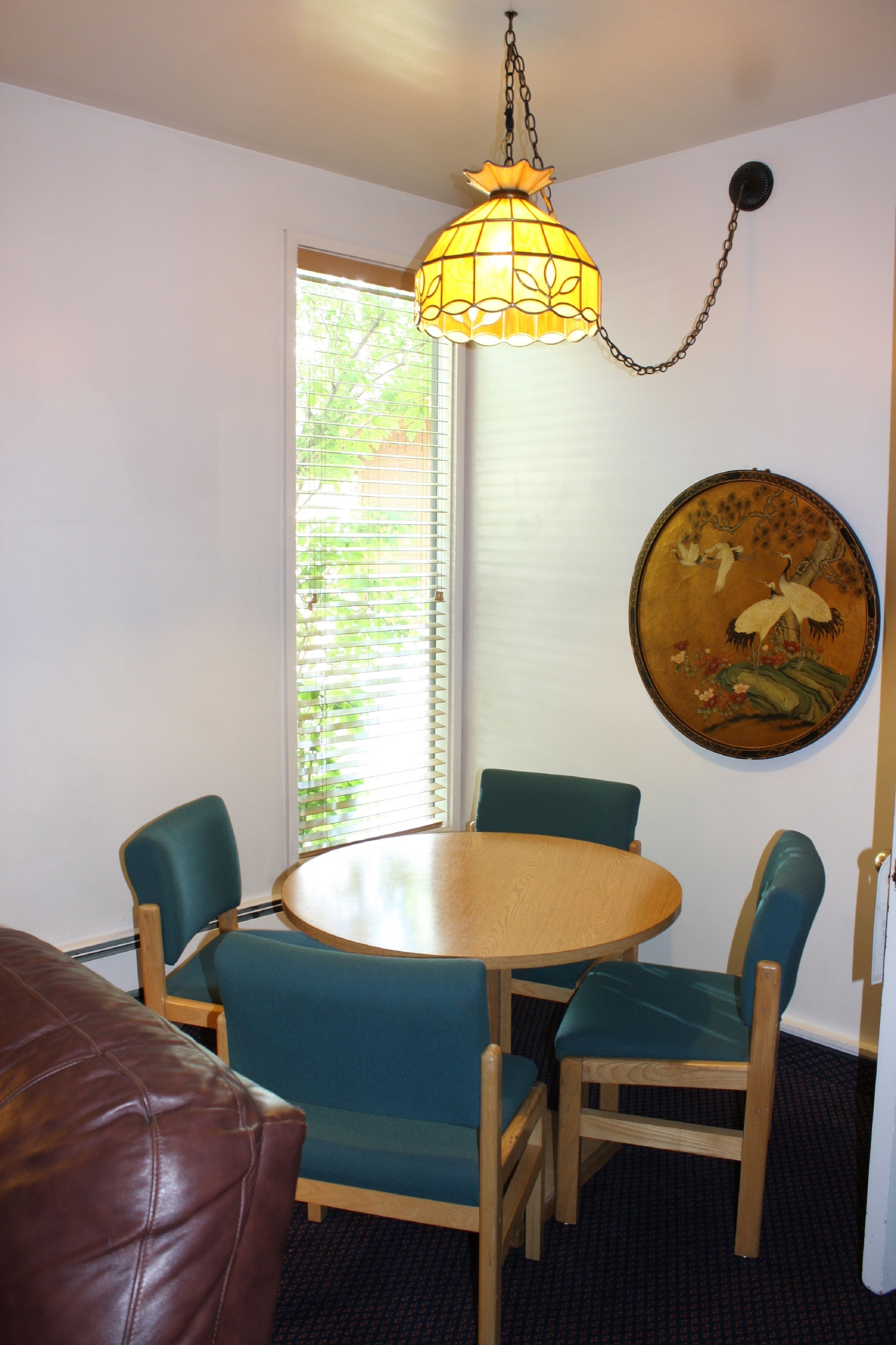 A dining area with a round wooden table, four chairs, a hanging light fixture, a window with blinds, and a wall-mounted painting.