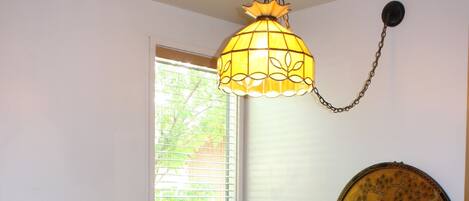 A dining area with a round wooden table, four chairs, a hanging light fixture, a window with blinds, and a wall-mounted painting.