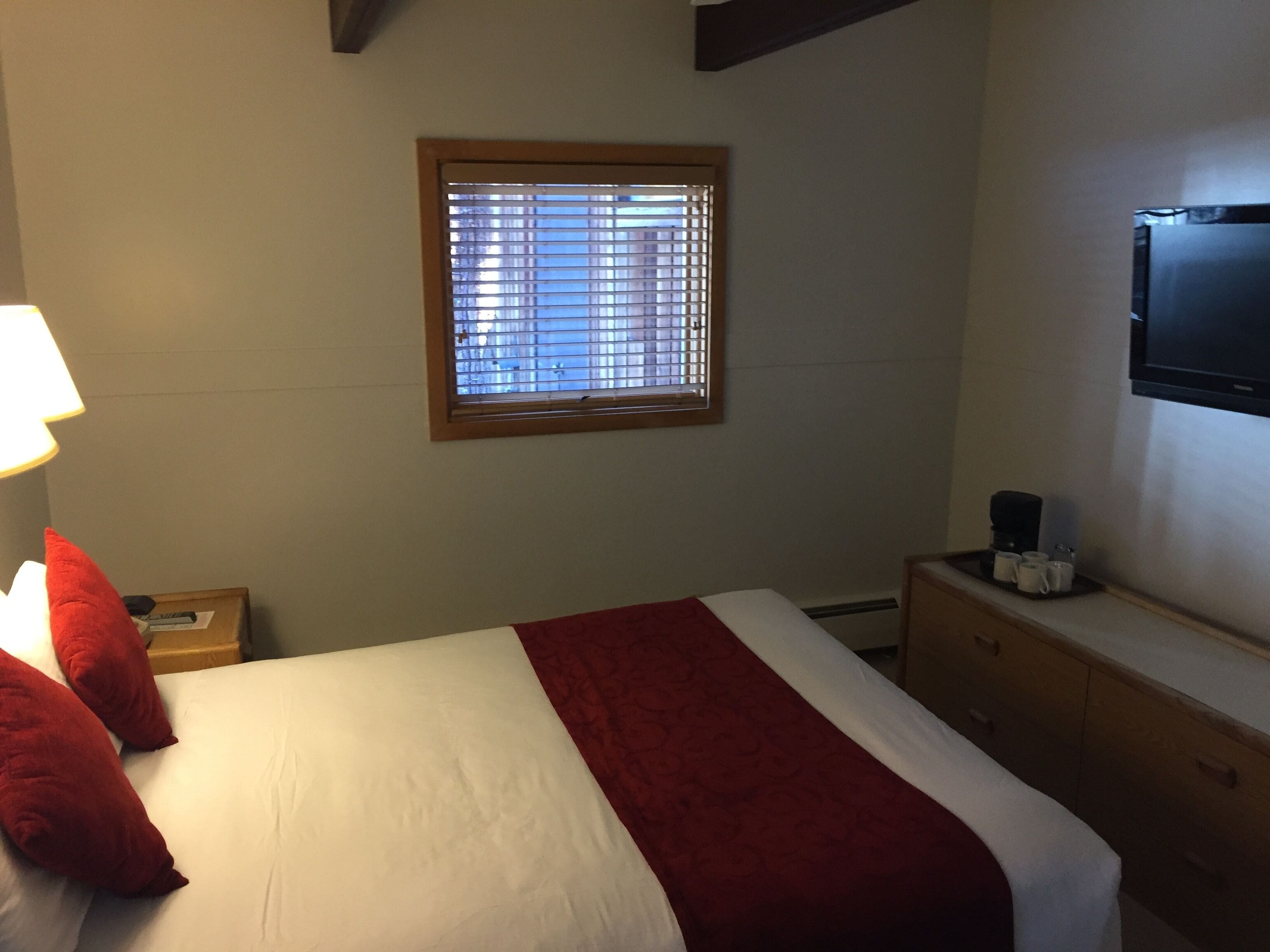 Una habitación de hotel con una cama, un ventanal con persianas, un televisor montado en la pared y una cafetera sobre una mesita.
