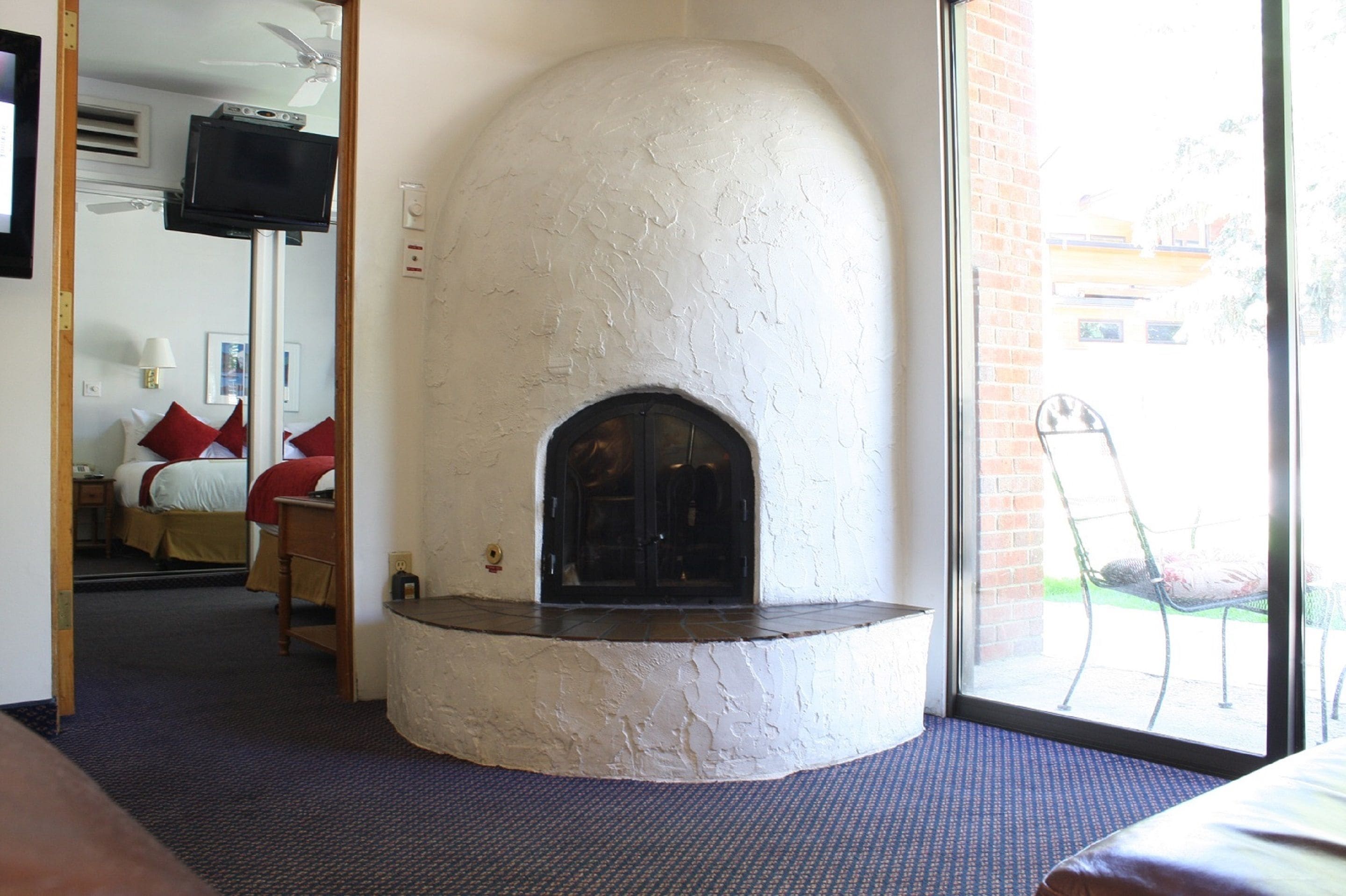 A room with a fireplace, a bed with red pillows, a TV mounted on a wall, and a window with a view of the outside.