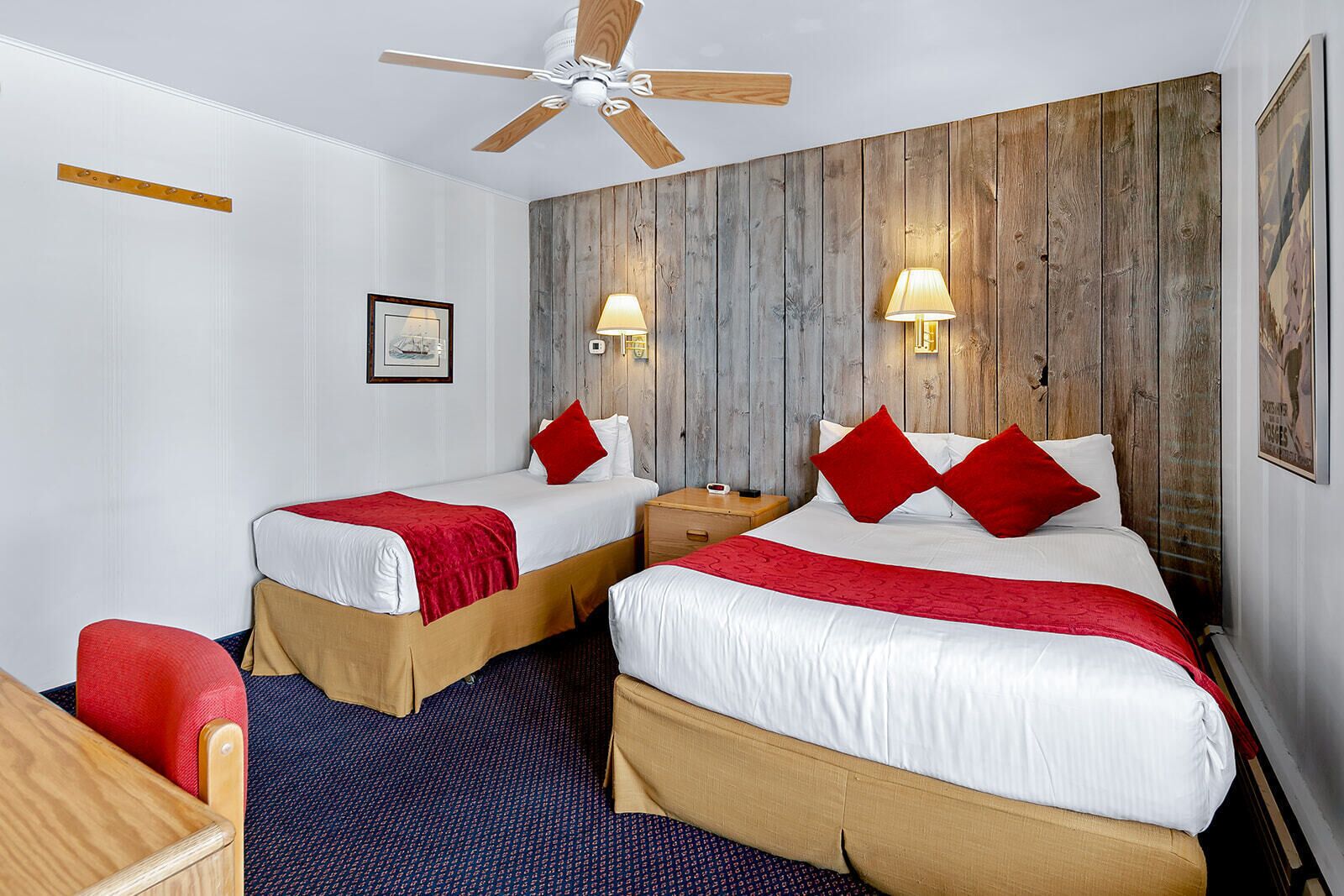 Une chambre d’hôtel avec deux lits, des panneaux en bois au niveau de la tête de lit, un ventilateur de plafond et une chaise rouge.