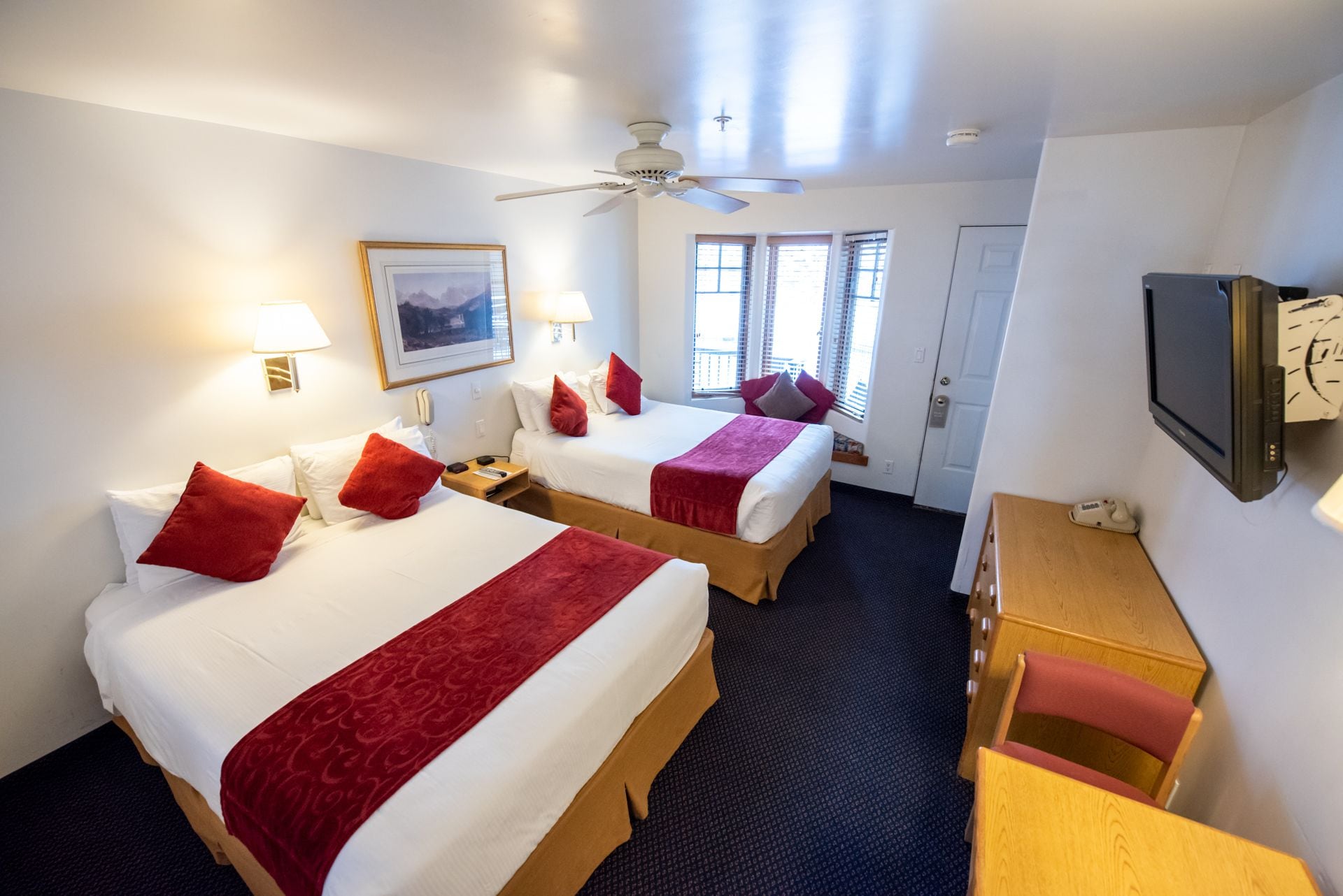 Une chambre d’hôtel avec deux lits, un bureau, une chaise, une télévision et un ventilateur de plafond.